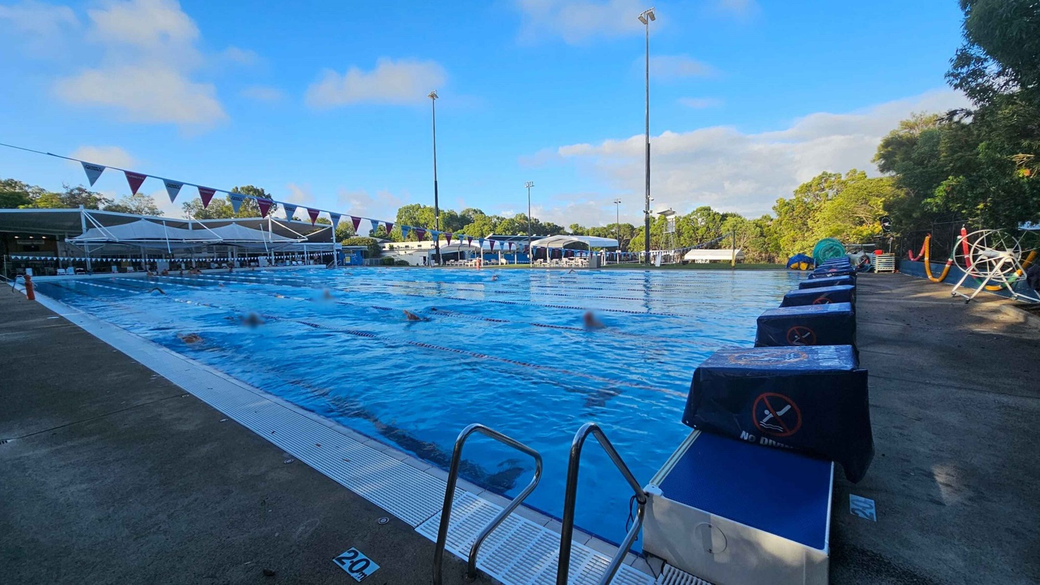 Outdoor swimming pool with several lanes, surrounded by a concrete deck. On the left, there are shaded seating areas with white canopies. Swimmers are in the pool, and lane dividers are visible. On the right, there are starting blocks covered with blue fabric and a lifeguard chair. Trees and greenery are in the background, with tall floodlights around the pool area. Outdoor swimming pool with several lanes, surrounded by a concrete deck. On the left, there are shaded seating areas with white canopies. Swimmers are in the pool, and lane dividers are visible. On the right, there are starting blocks covered with blue fabric and a lifeguard chair. Trees and greenery are in the background, with tall floodlights around the pool area.