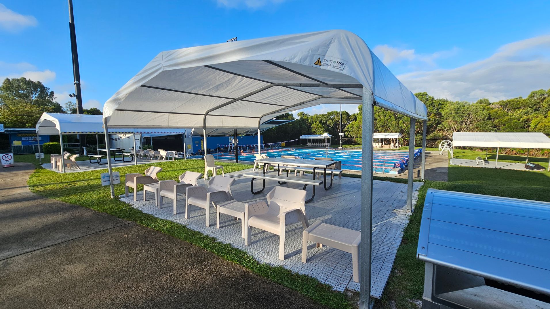 A swimming pool area with a large white canopy in the foreground, covering several white plastic chairs and picnic tables. To the left, another canopy shelters more seating. The pool, with lane dividers, is visible in the middle distance, surrounded by green grass and trees. A paved path runs along the left side, and a blue structure is partially visible in the background. A swimming pool area with a large white canopy in the foreground, covering several white plastic chairs and picnic tables. To the left, another canopy shelters more seating. The pool, with lane dividers, is visible in the middle distance, surrounded by green grass and trees. A paved path runs along the left side, and a blue structure is partially visible in the background.