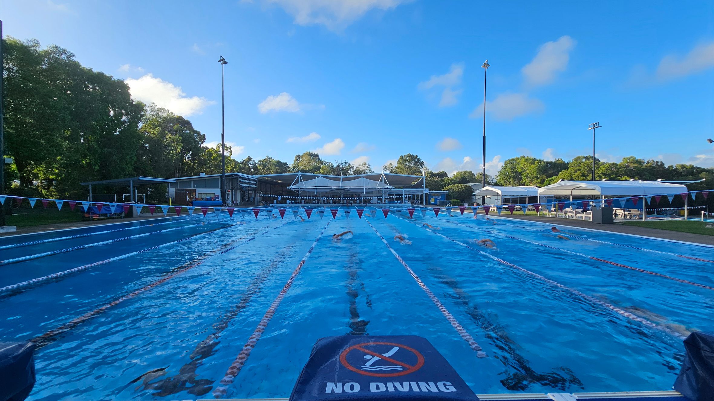 An outdoor swimming pool with several lanes, each marked by lane dividers. Swimmers are actively swimming in the lanes. A "No Diving" sign is visible in the foreground. On the left, there are trees and a small building. On the right, there are white tents and chairs. In the background, more trees and a building with a white roof are visible. The pool area is well-lit with tall floodlights. An outdoor swimming pool with several lanes, each marked by lane dividers. Swimmers are actively swimming in the lanes. A "No Diving" sign is visible in the foreground. On the left, there are trees and a small building. On the right, there are white tents and chairs. In the background, more trees and a building with a white roof are visible. The pool area is well-lit with tall floodlights.