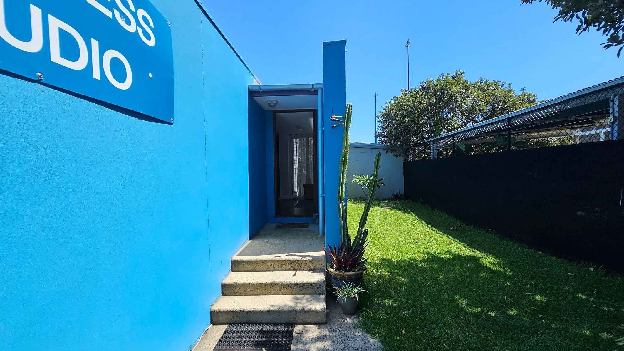 A bright blue building with a sign partially visible on the left. In the centre, a small set of concrete steps leads to a glass door. To the right, a tall cactus and other potted plants are placed on a concrete surface. A grassy area extends to the right, bordered by a black fence and a metal structure. Trees are visible in the background. A bright blue building with a sign partially visible on the left. In the centre, a small set of concrete steps leads to a glass door. To the right, a tall cactus and other potted plants are placed on a concrete surface. A grassy area extends to the right, bordered by a black fence and a metal structure. Trees are visible in the background.