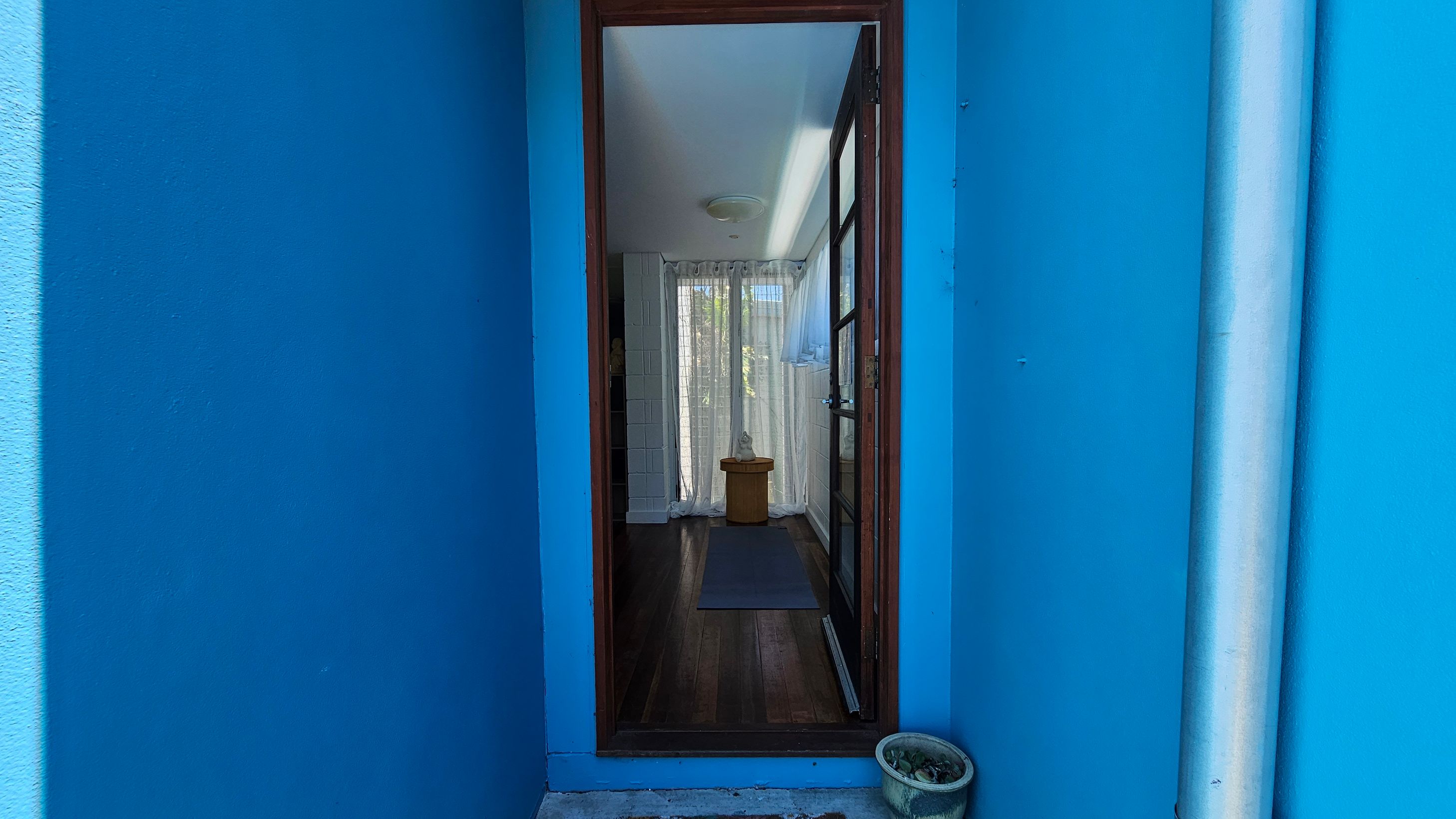 A narrow entrance with bright blue walls on either side leads to a wooden door with glass panels. Inside, there is a wooden floor and a small table with a decorative item on it. Sheer curtains cover a window at the back, allowing light to filter through. A small potted plant is on the ground outside, near the entrance. A white ceiling is visible inside the room. A narrow entrance with bright blue walls on either side leads to a wooden door with glass panels. Inside, there is a wooden floor and a small table with a decorative item on it. Sheer curtains cover a window at the back, allowing light to filter through. A small potted plant is on the ground outside, near the entrance. A white ceiling is visible inside the room.