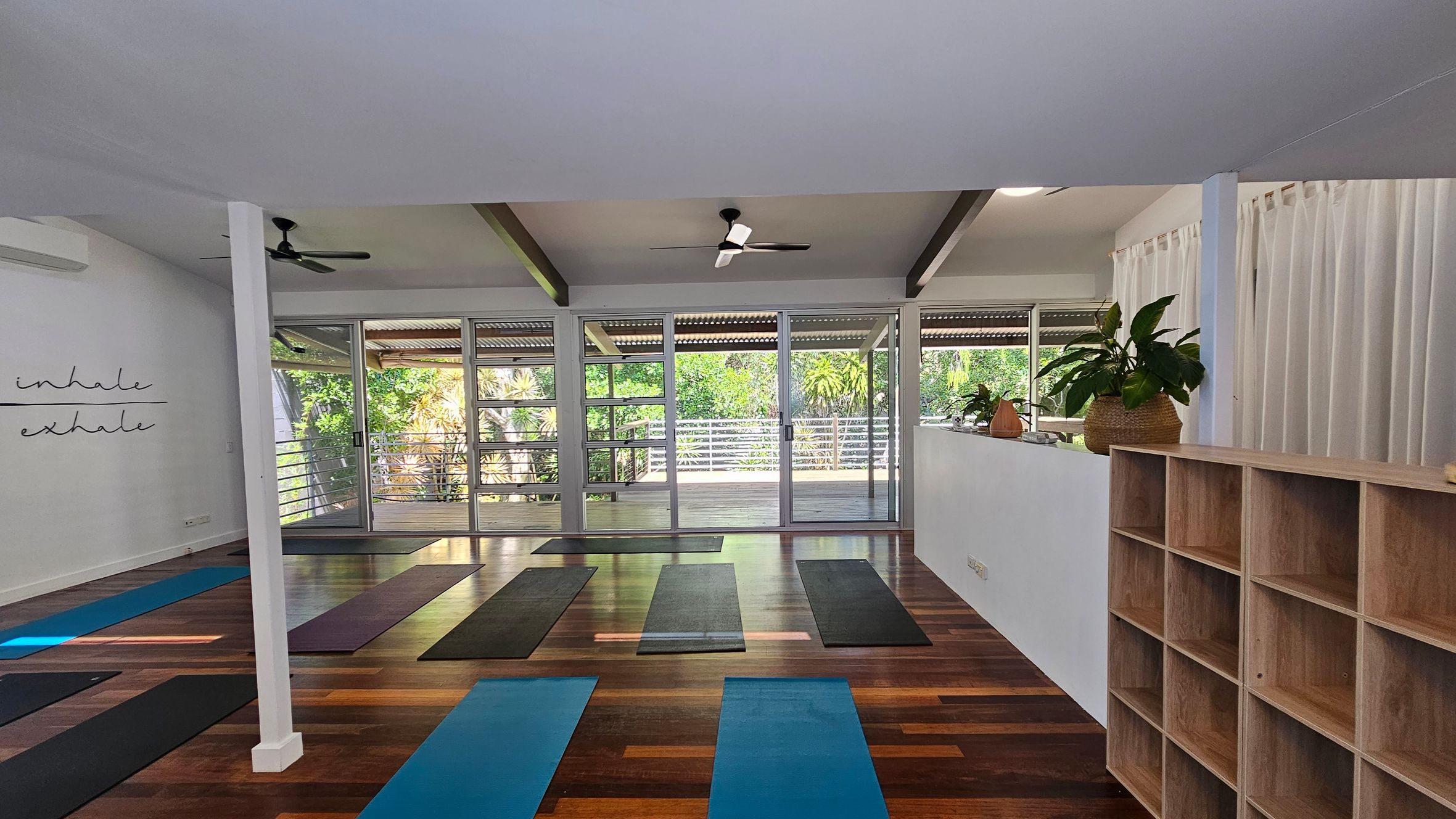 A spacious yoga studio with wooden flooring and a white ceiling. On the left, a wall displays the words "inhale" and "exhale." In the centre, several yoga mats are laid out in rows. The back wall features large glass doors and windows, offering a view of lush greenery outside. On the right, there is a wooden shelving unit and a ledge with potted plants. Ceiling fans are mounted above for ventilation. A spacious yoga studio with wooden flooring and a white ceiling. On the left, a wall displays the words "inhale" and "exhale." In the centre, several yoga mats are laid out in rows. The back wall features large glass doors and windows, offering a view of lush greenery outside. On the right, there is a wooden shelving unit and a ledge with potted plants. Ceiling fans are mounted above for ventilation.