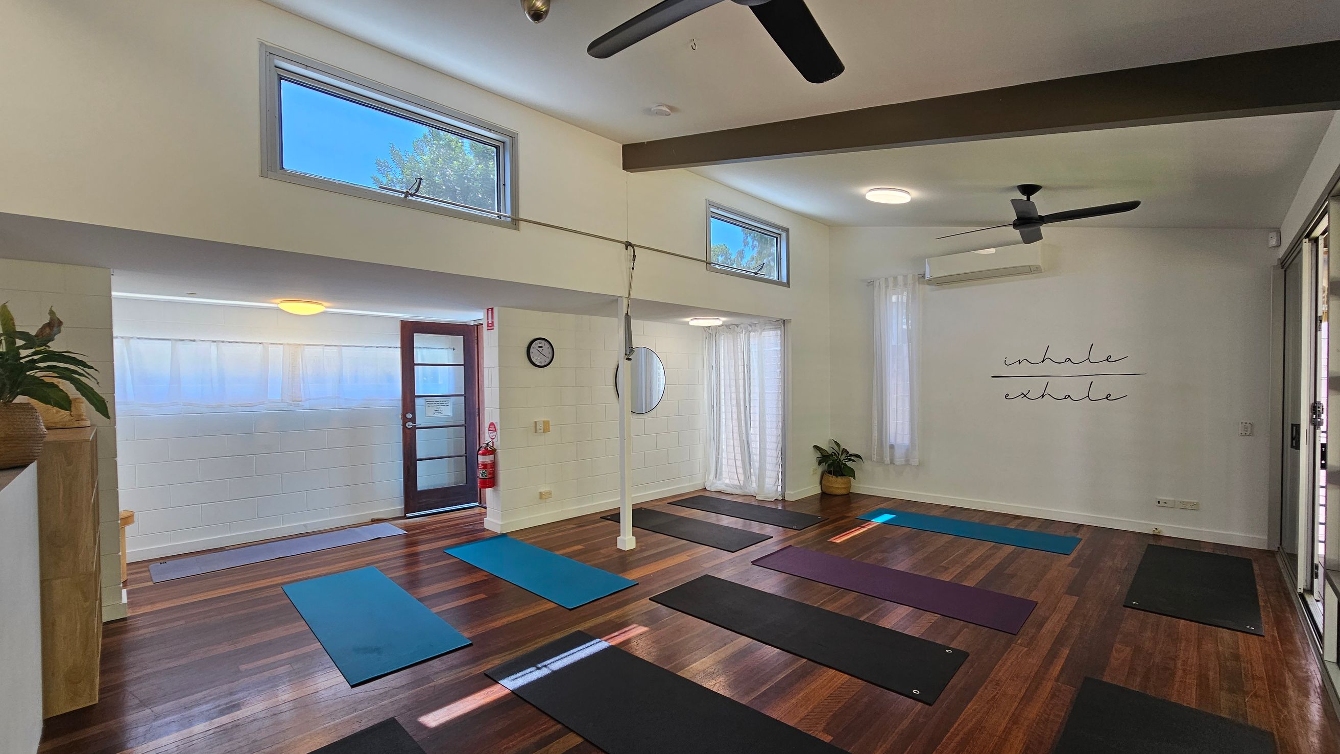 A yoga studio with wooden flooring and white walls. On the right, there are several yoga mats laid out in rows. The wall has the words "inhale" and "exhale" written on it. On the left, a door with frosted glass panels is visible, along with a clock and a circular mirror on the wall. There are ceiling fans and high windows allowing natural light. A potted plant is in the corner, and a fire extinguisher is near the door. A yoga studio with wooden flooring and white walls. On the right, there are several yoga mats laid out in rows. The wall has the words "inhale" and "exhale" written on it. On the left, a door with frosted glass panels is visible, along with a clock and a circular mirror on the wall. There are ceiling fans and high windows allowing natural light. A potted plant is in the corner, and a fire extinguisher is near the door.