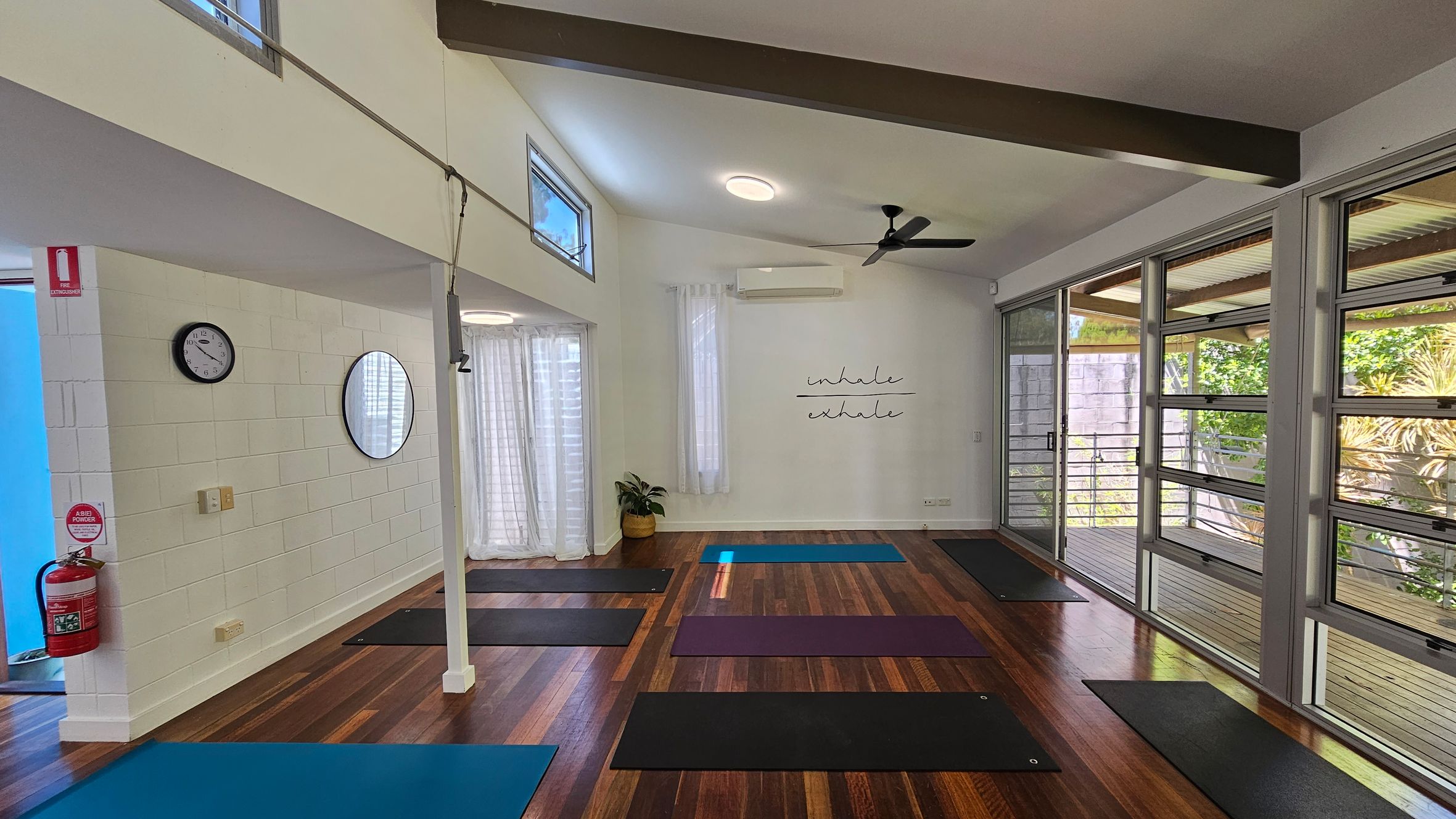 A yoga studio with wooden flooring and a high ceiling. On the left, a white brick wall features a clock, a round mirror, and a fire extinguisher. Yoga mats are spread across the floor. The back wall has a plant and a sign reading "inhale, exhale." Large windows on the right let in natural light, with views of greenery outside. Sheer curtains hang by the windows, and a ceiling fan is visible above. A yoga studio with wooden flooring and a high ceiling. On the left, a white brick wall features a clock, a round mirror, and a fire extinguisher. Yoga mats are spread across the floor. The back wall has a plant and a sign reading "inhale, exhale." Large windows on the right let in natural light, with views of greenery outside. Sheer curtains hang by the windows, and a ceiling fan is visible above.