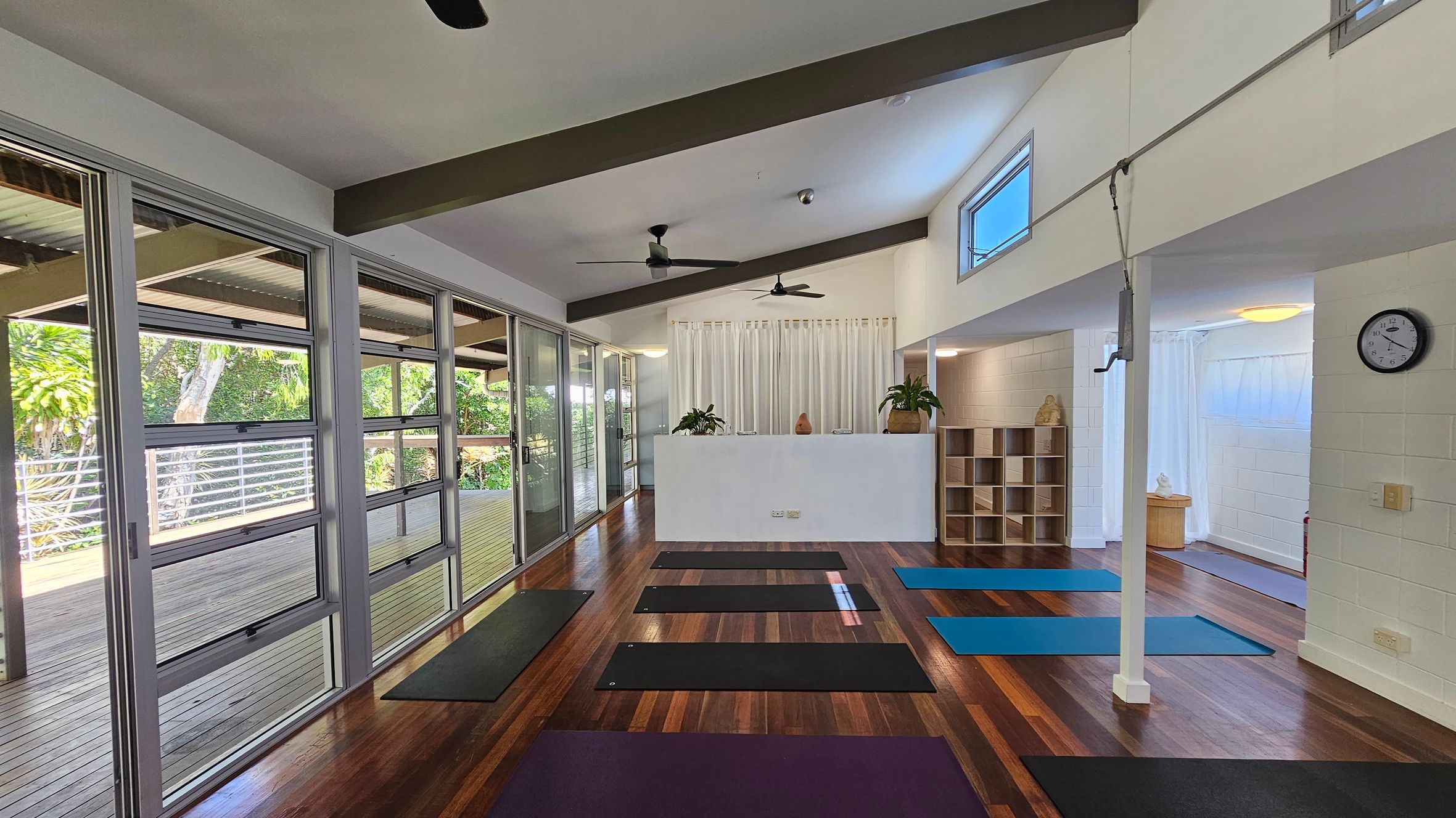A yoga studio with wooden flooring and several yoga mats laid out in rows. On the left, large glass windows and doors open to a wooden deck with greenery outside. In the middle, a white partition with plants on top separates the space. On the right, a wall clock is mounted above a shelf unit with cubbyholes. The ceiling has exposed beams and ceiling fans. A yoga studio with wooden flooring and several yoga mats laid out in rows. On the left, large glass windows and doors open to a wooden deck with greenery outside. In the middle, a white partition with plants on top separates the space. On the right, a wall clock is mounted above a shelf unit with cubbyholes. The ceiling has exposed beams and ceiling fans.