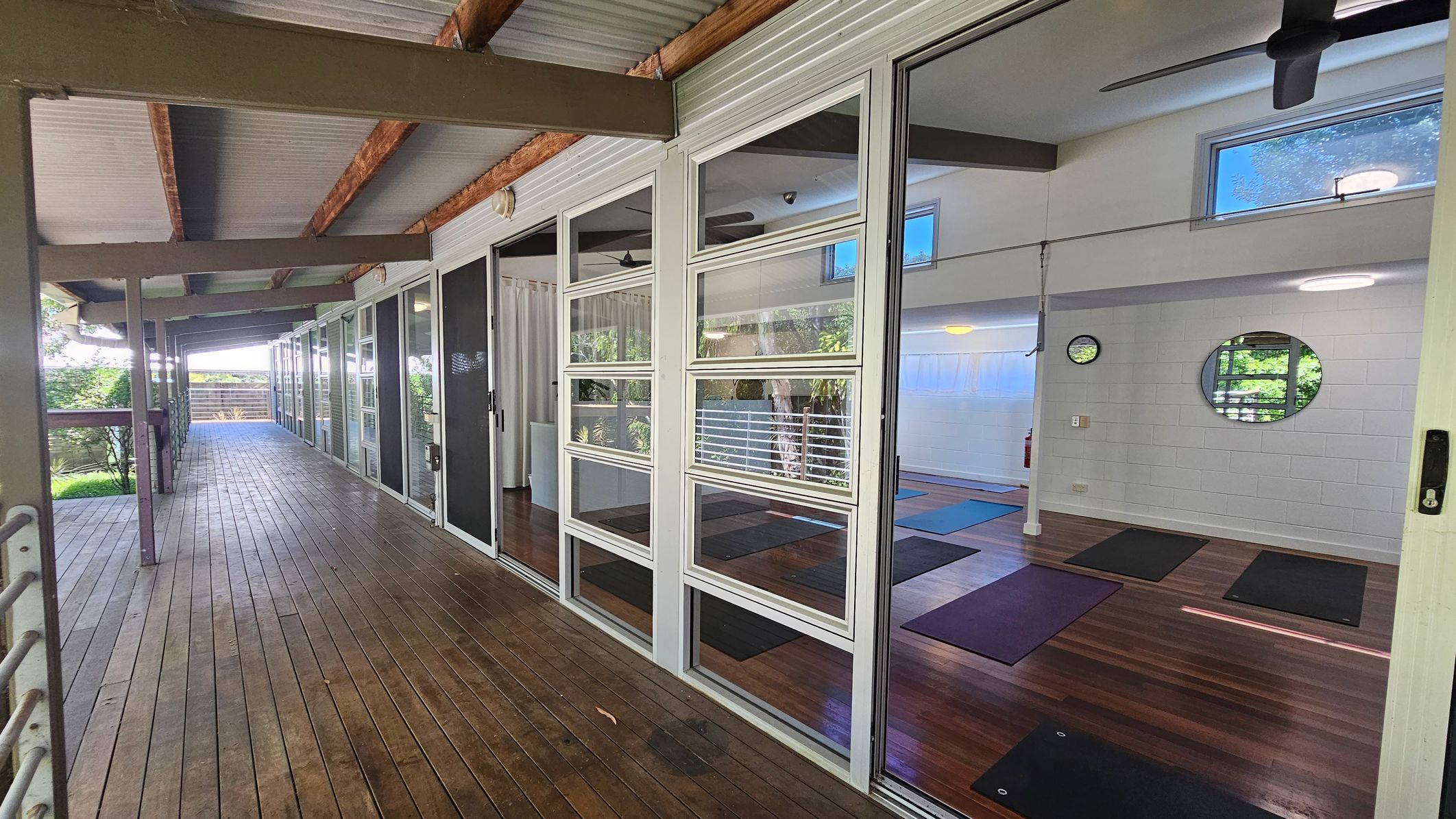 A wooden deck runs along the left side, bordered by a railing and lush greenery. On the right, large glass doors reveal an interior room with yoga mats on a wooden floor. The room has white walls, a circular mirror, and a clock. The ceiling is high with exposed beams and ceiling fans. A wooden deck runs along the left side, bordered by a railing and lush greenery. On the right, large glass doors reveal an interior room with yoga mats on a wooden floor. The room has white walls, a circular mirror, and a clock. The ceiling is high with exposed beams and ceiling fans.