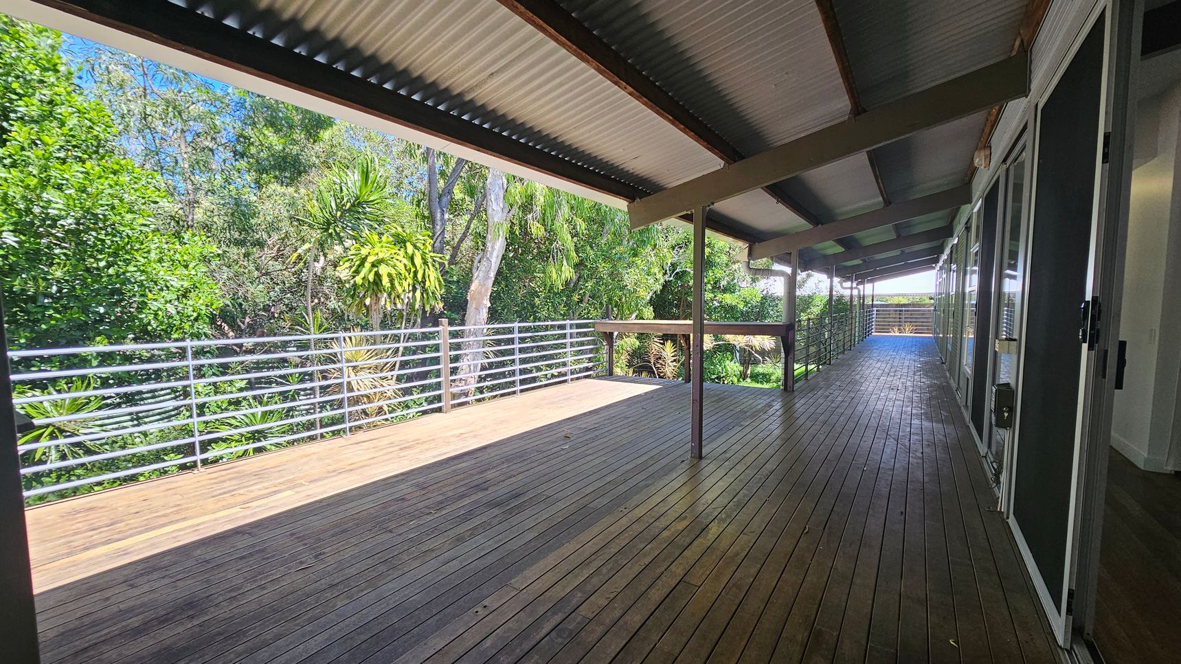 A wooden deck with a corrugated metal roof extends along the right side, where glass doors are visible. On the left, a metal railing borders the deck, overlooking lush green foliage and trees. The floor is made of wooden planks, and the ceiling is supported by beams. The deck is spacious and open, providing a view of the surrounding greenery. A wooden deck with a corrugated metal roof extends along the right side, where glass doors are visible. On the left, a metal railing borders the deck, overlooking lush green foliage and trees. The floor is made of wooden planks, and the ceiling is supported by beams. The deck is spacious and open, providing a view of the surrounding greenery.