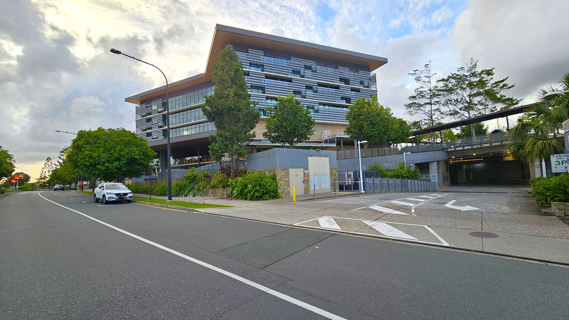 A modern multi-storey building with a flat roof is on the right, featuring horizontal lines and large windows. In front, there are several trees and a landscaped area with shrubs. A road runs along the left side, with a white car parked on the street. The pavement is marked with arrows and parking signs. A covered entrance to a car park is visible on the right, with greenery surrounding the area. Streetlights line the road, and the sky is overcast.