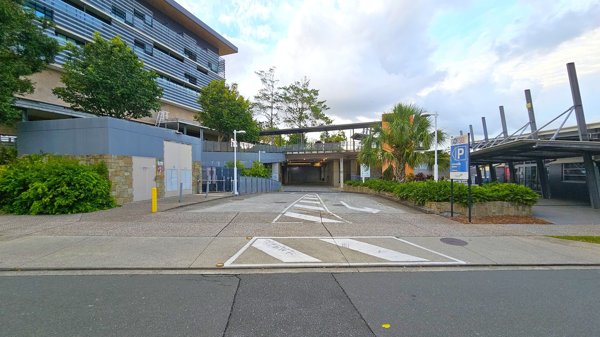 A modern building with horizontal striped cladding on the left, surrounded by trees and shrubs. In the centre, a driveway leads to an underground parking area, with directional arrows painted on the ground. On the right, a covered walkway with a blue parking sign and more greenery. The foreground features a paved area with a mix of concrete and grass. The background includes a few tall trees and a partly cloudy sky.
