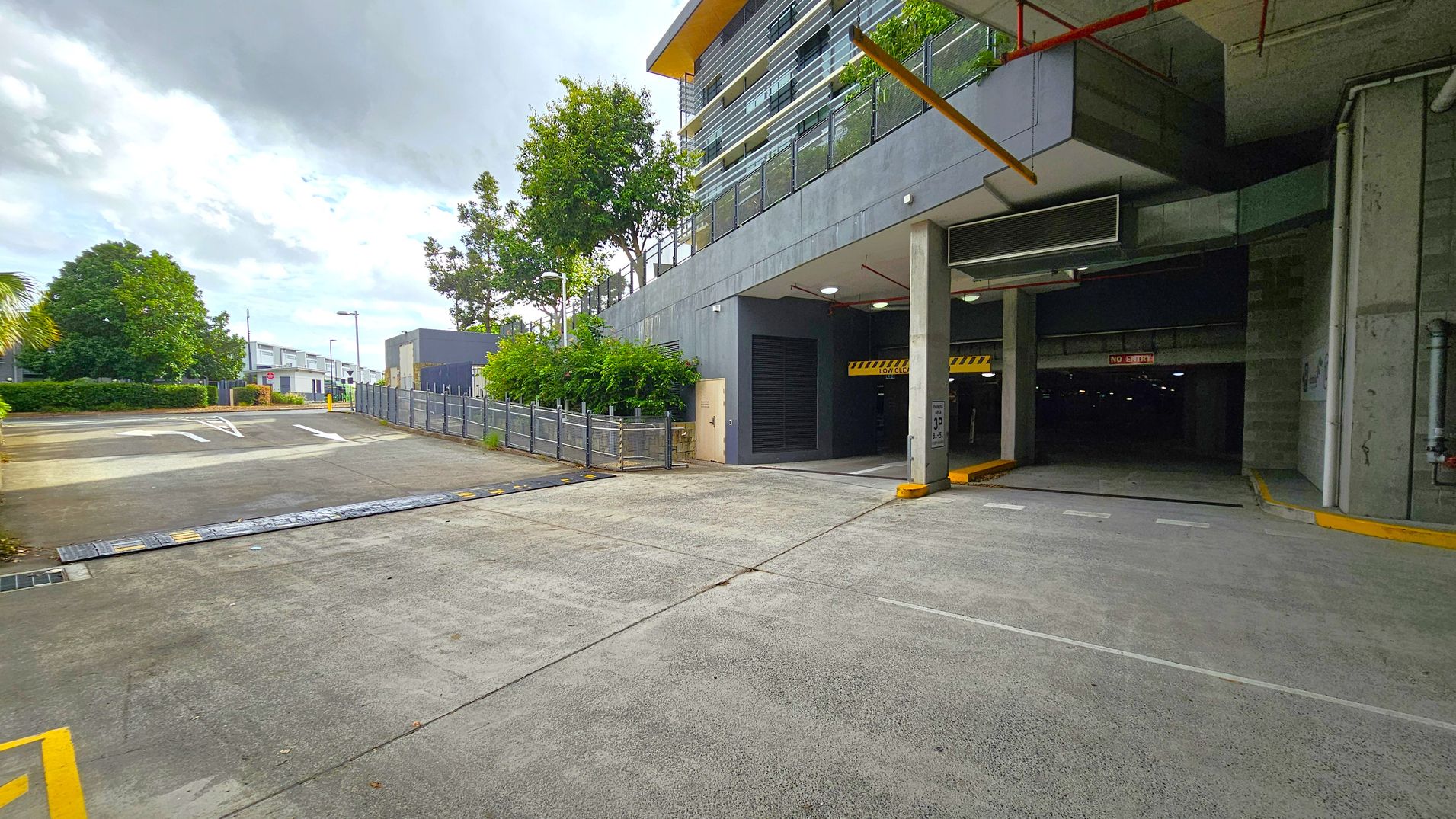 A concrete driveway leads to an underground car park on the right, marked by a "No Entry" sign. The building above has a modern design with horizontal lines and a yellow roof edge. On the left, a fenced area with trees and shrubs lines the path, leading to a small building in the distance. The ground is paved, and the ceiling of the car park entrance has visible pipes and lighting.