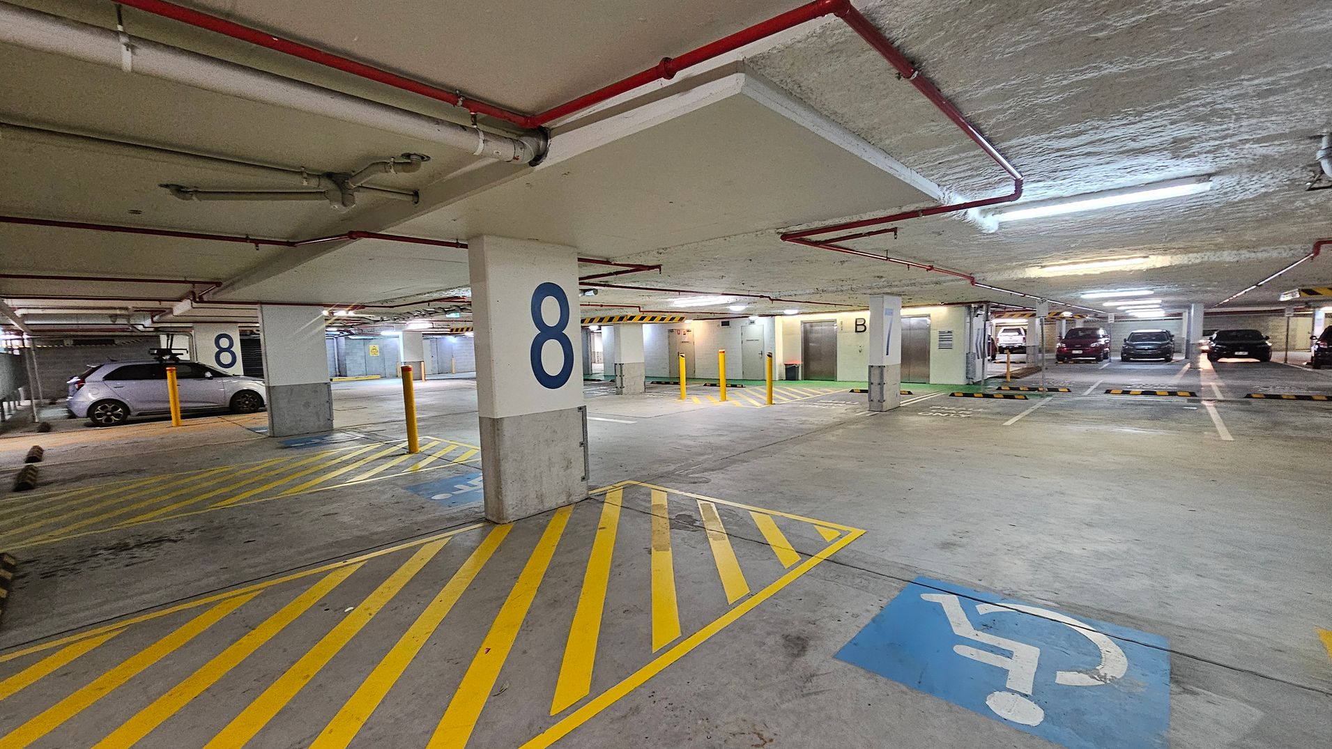 A basement car park with a concrete floor and ceiling. On the left, a silver car is parked next to a pillar marked with the number 8. Yellow diagonal lines mark the ground near the pillar. In the foreground, a blue disabled parking symbol is visible. The right side shows several parked cars in marked bays. The ceiling has exposed pipes and fluorescent lighting. Yellow bollards are positioned near the centre, and a sign with the letter B is visible on another pillar.