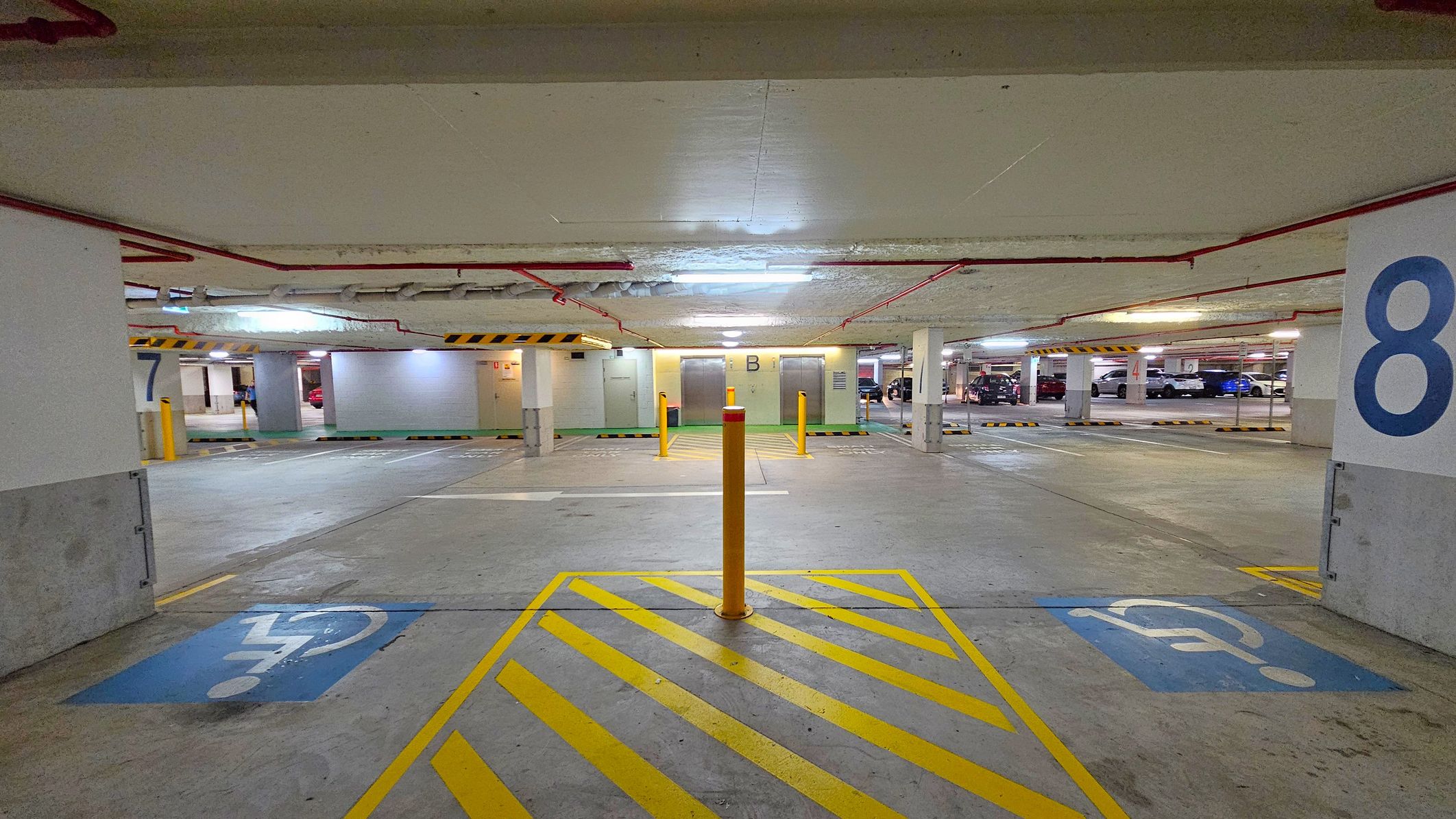 Underground car park with a concrete floor and ceiling. In the foreground, there are yellow diagonal lines and a bollard, with blue disabled parking symbols on either side. The background shows parking spaces numbered 7 and 8 on the left and right, respectively. Two lifts marked "B" are in the centre, with cars parked further back. Red pipes run along the ceiling, and fluorescent lights illuminate the area.