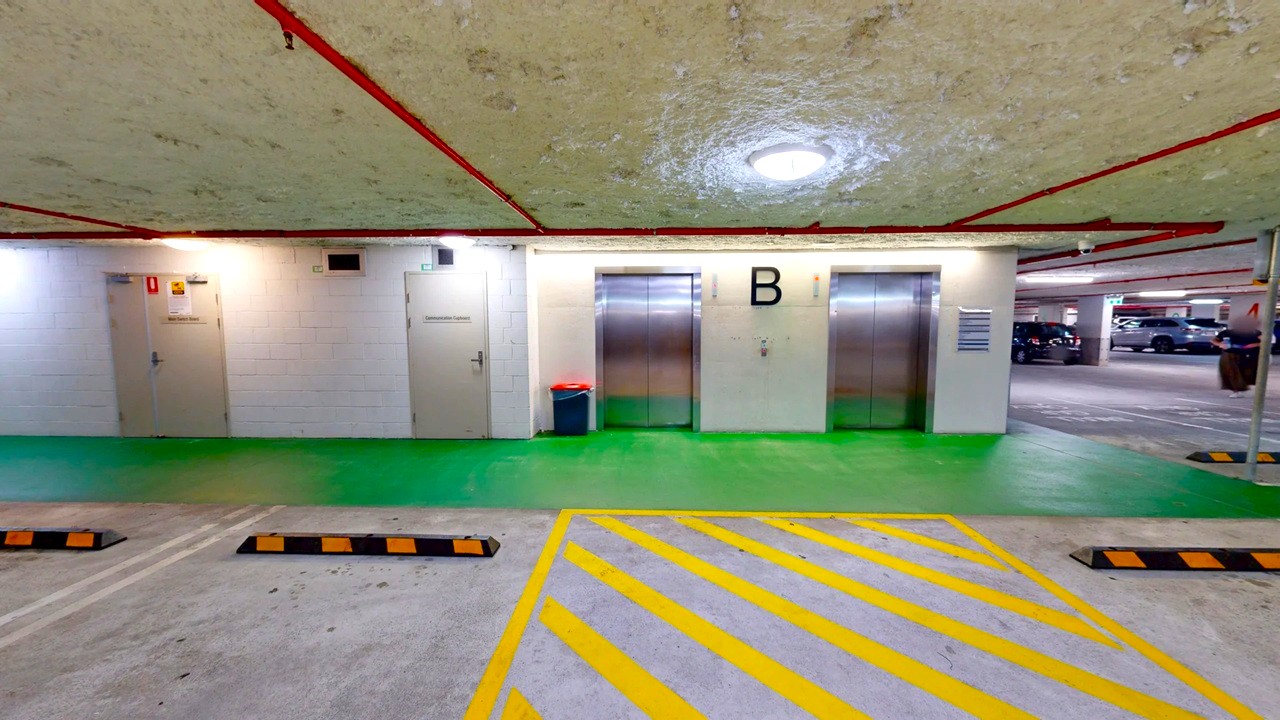 In an underground car park, two metallic lift doors are centred on a white wall, marked with a large letter "B". To the left, two grey doors are visible, with a red bin nearby. The floor is a mix of green and grey, with yellow diagonal lines in the foreground. The ceiling is textured with red pipes running across it. On the right, parked cars are partially visible.