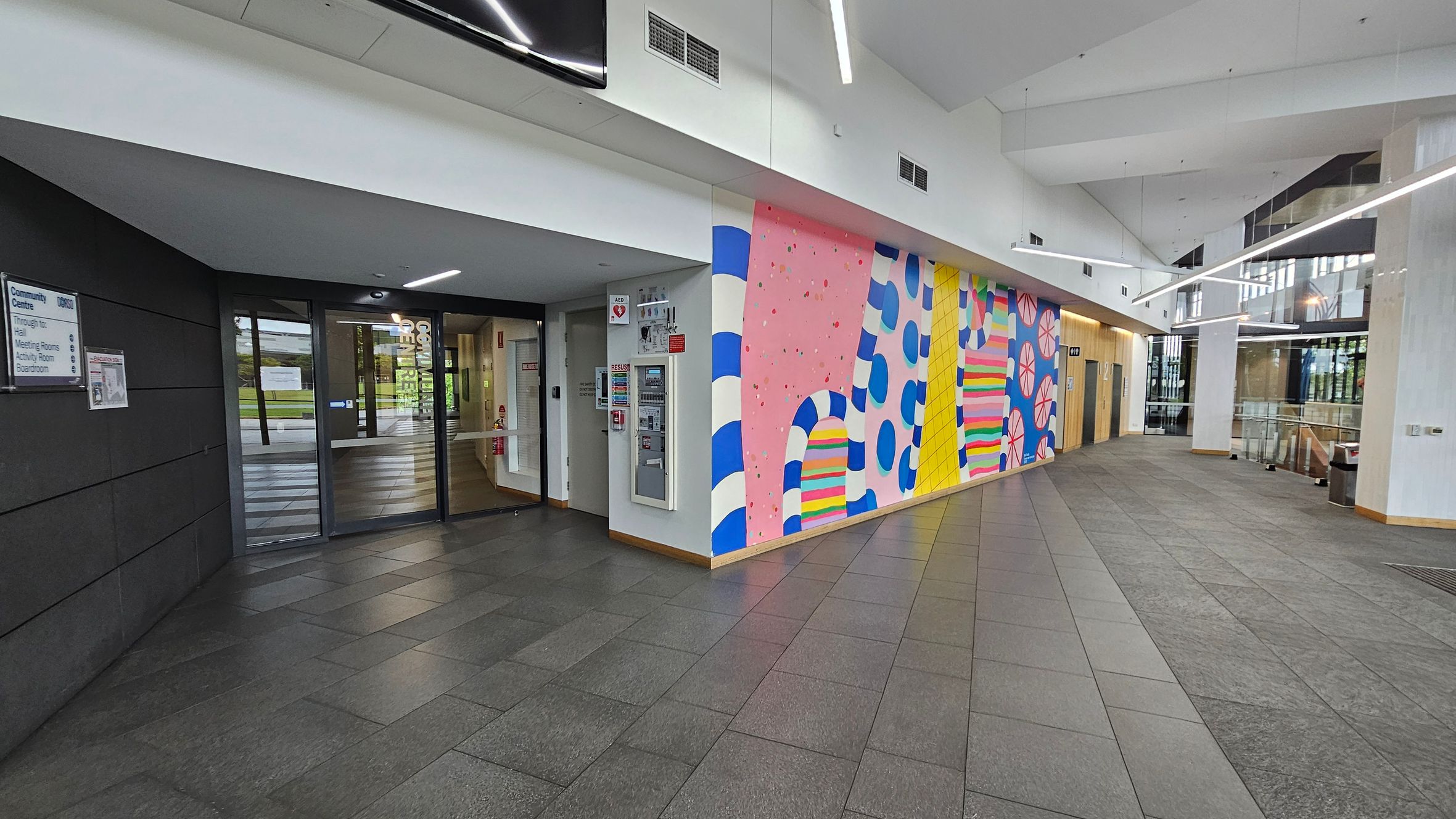 A spacious indoor area with a dark tiled floor. On the left, there's a glass entrance with a sign indicating a community centre and various rooms. The wall next to the entrance features a colourful mural with abstract patterns in pink, blue, yellow, and green. On the right, the space opens up with large windows allowing natural light. The ceiling is white with modern linear lighting fixtures.