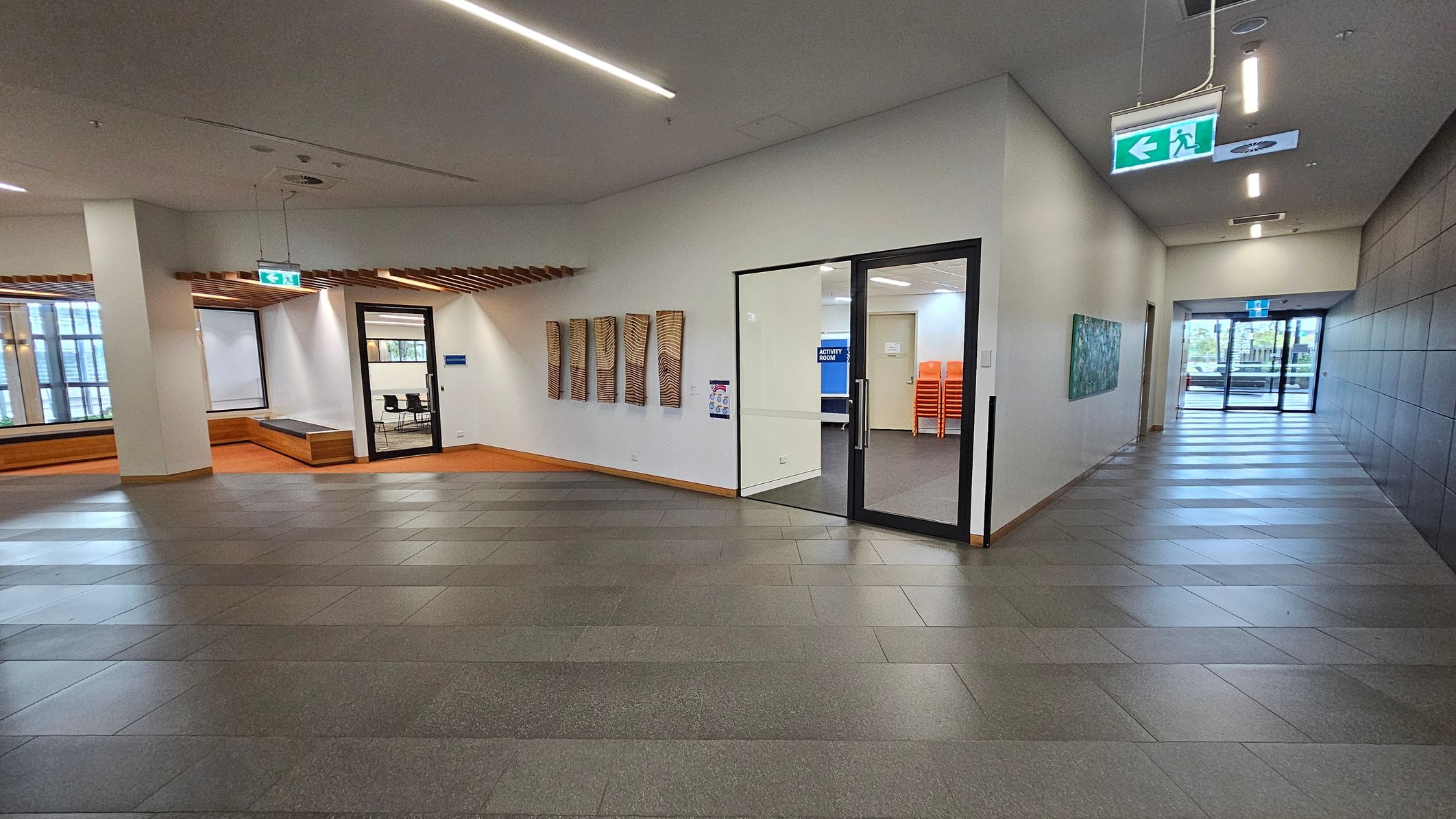 A modern indoor space with a tiled floor and a white ceiling featuring linear lights. On the left, there's a seating area with wooden benches and a large window. In the centre, a glass door leads to a room labelled "Activity Room," with stacked orange chairs visible inside. The right side has a corridor with a green exit sign and a piece of artwork on the wall. The background shows glass doors leading outside.