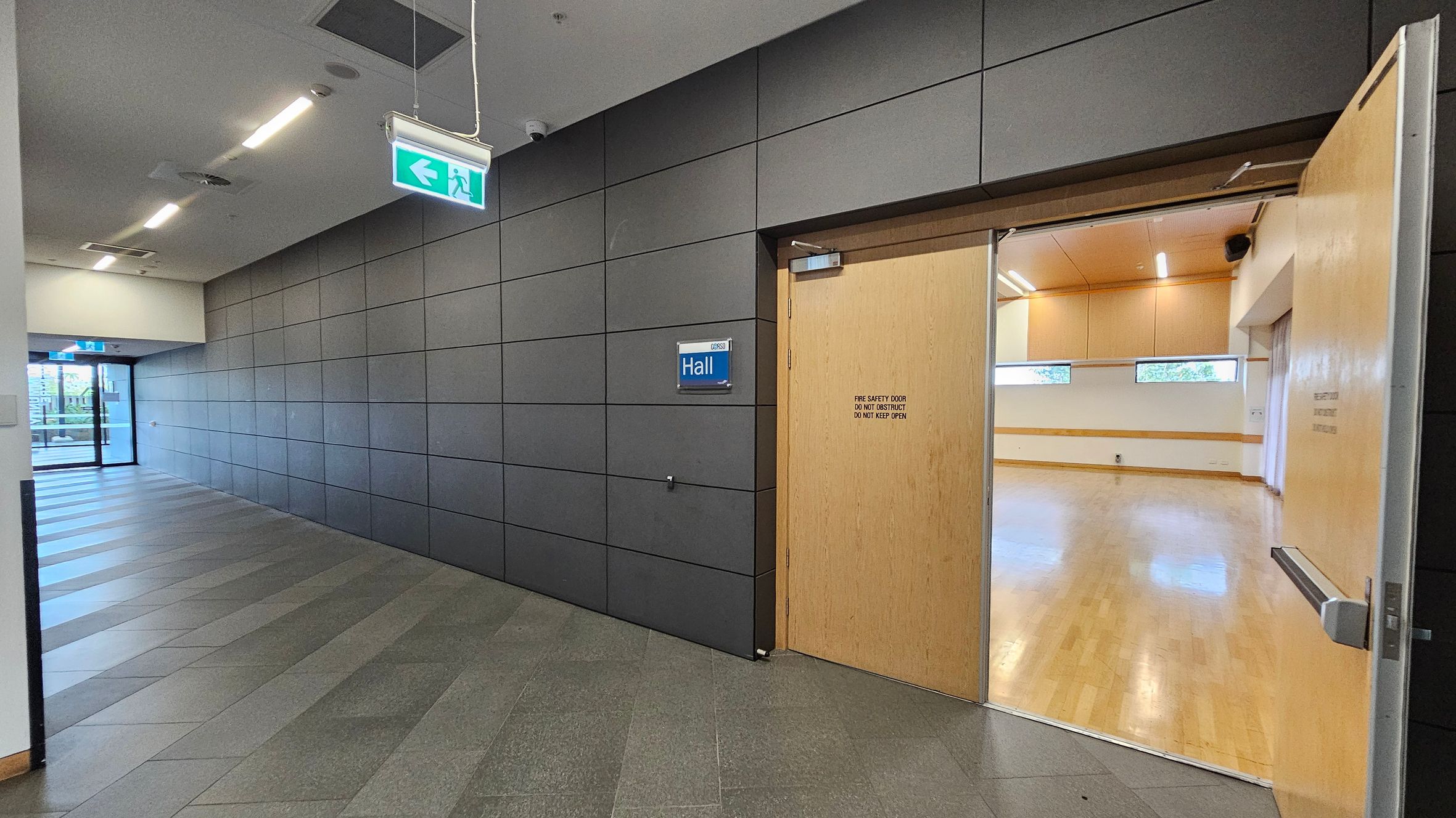 A hallway with grey tiled walls and a striped floor leads to a glass door at the end. On the right, a wooden door marked "Hall" is open, revealing a room with light wooden flooring and small rectangular windows. The ceiling has recessed lighting, and an illuminated exit sign hangs above the hallway.