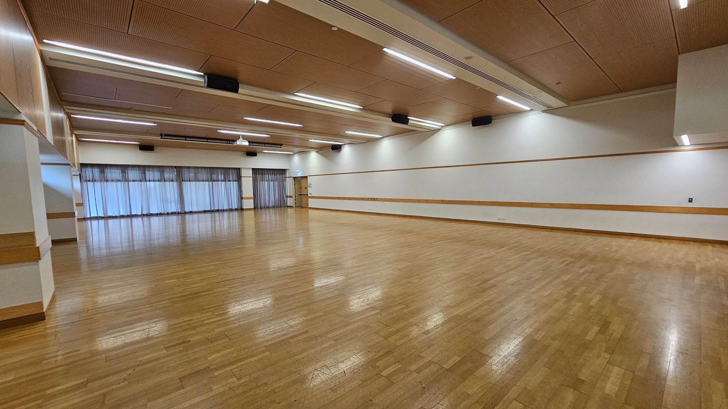 A spacious room with a polished wooden floor and a light brown ceiling featuring recessed lighting. The left side has large windows covered with sheer curtains, allowing diffused light to enter. The right wall is plain with a wooden trim running horizontally. At the far end, there is a door and more windows. The room is empty, providing an open area suitable for various activities.