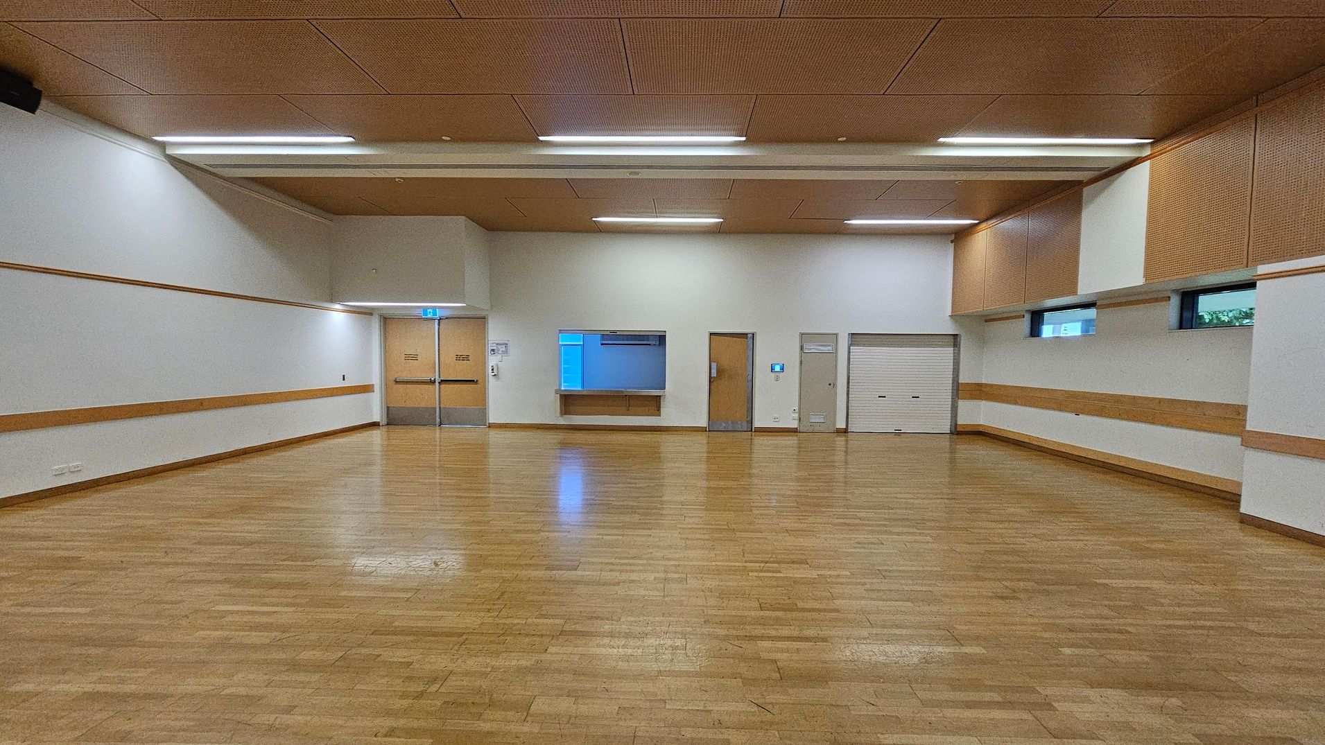 A spacious room with a wooden floor and a ceiling featuring recessed lighting. The walls are white with wooden trim. At the far end, there are double doors with a blue exit sign above, a service counter with a shutter, and a small window. To the right, there are two additional doors and a closed roller shutter. The room has high windows near the ceiling on the right side, allowing natural light to enter.