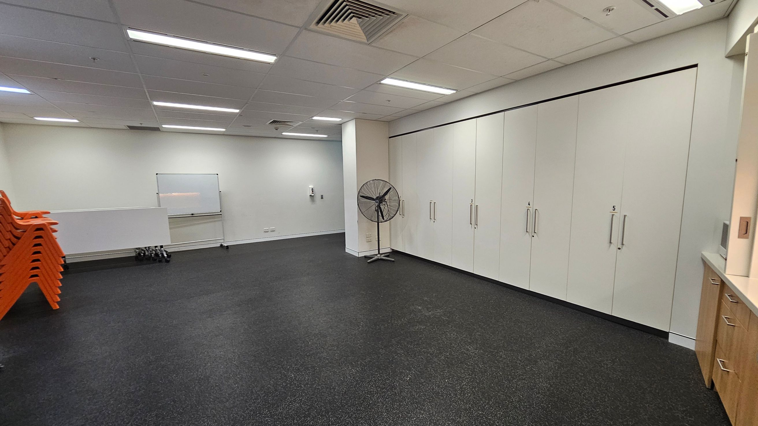 A room with a dark speckled floor and a white ceiling with fluorescent lights. On the left, there are stacks of orange chairs and a whiteboard on the wall. In the centre, a large fan stands near a column. On the right, there are light-coloured cabinets with handles, numbered 4 and 5, and a wooden cabinet with drawers. The walls are light-coloured, and there is a small set of dumbbells on the floor near the whiteboard.
