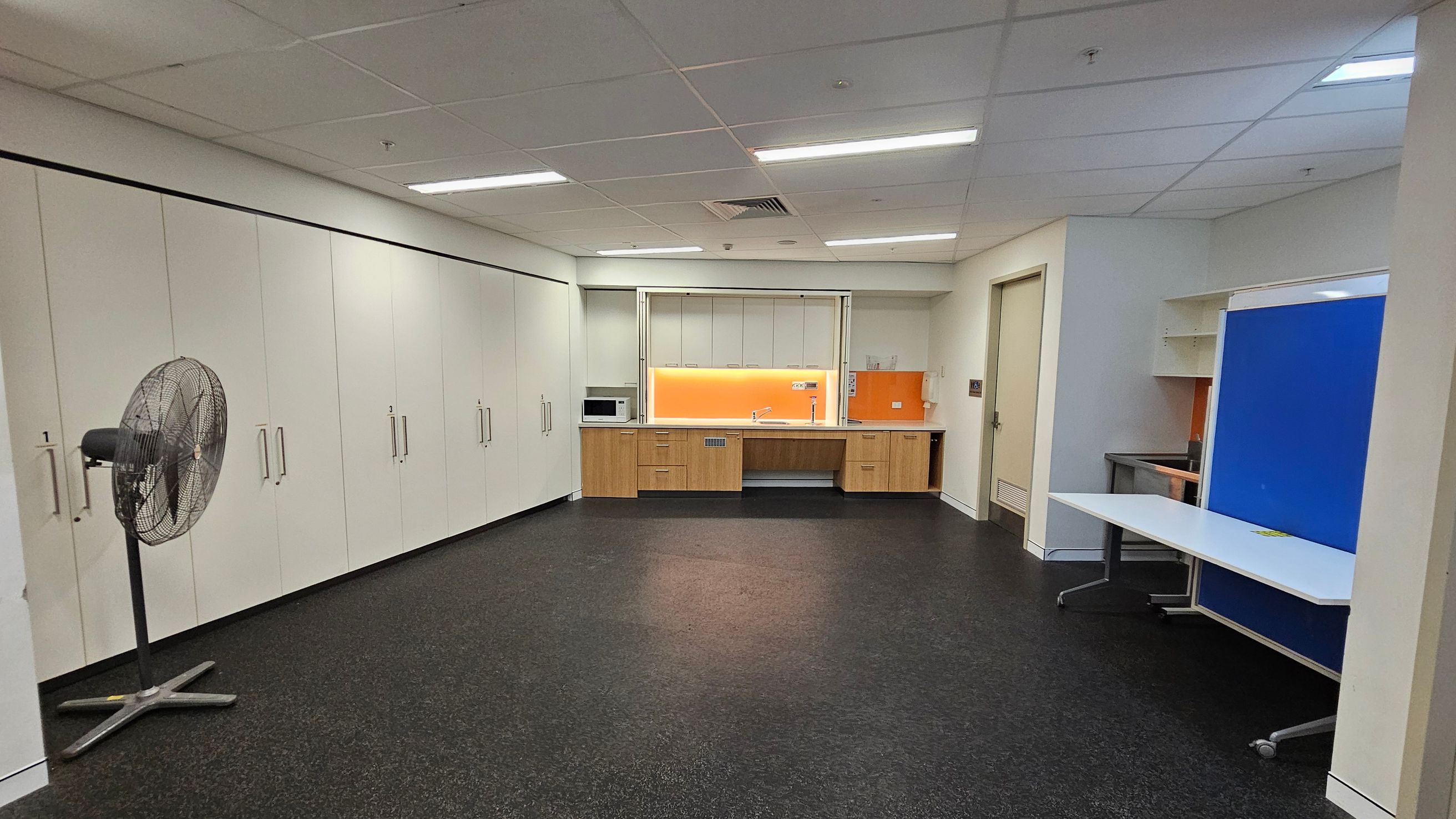 A room with a dark speckled floor and a white panelled ceiling with fluorescent lights. On the left, there is a large fan and a wall of white cabinets with handles. In the centre, a kitchenette features wooden cabinets, a microwave, and an orange splashback. On the right, a white table with wheels is positioned in front of a blue partition. A door is visible next to the kitchenette.