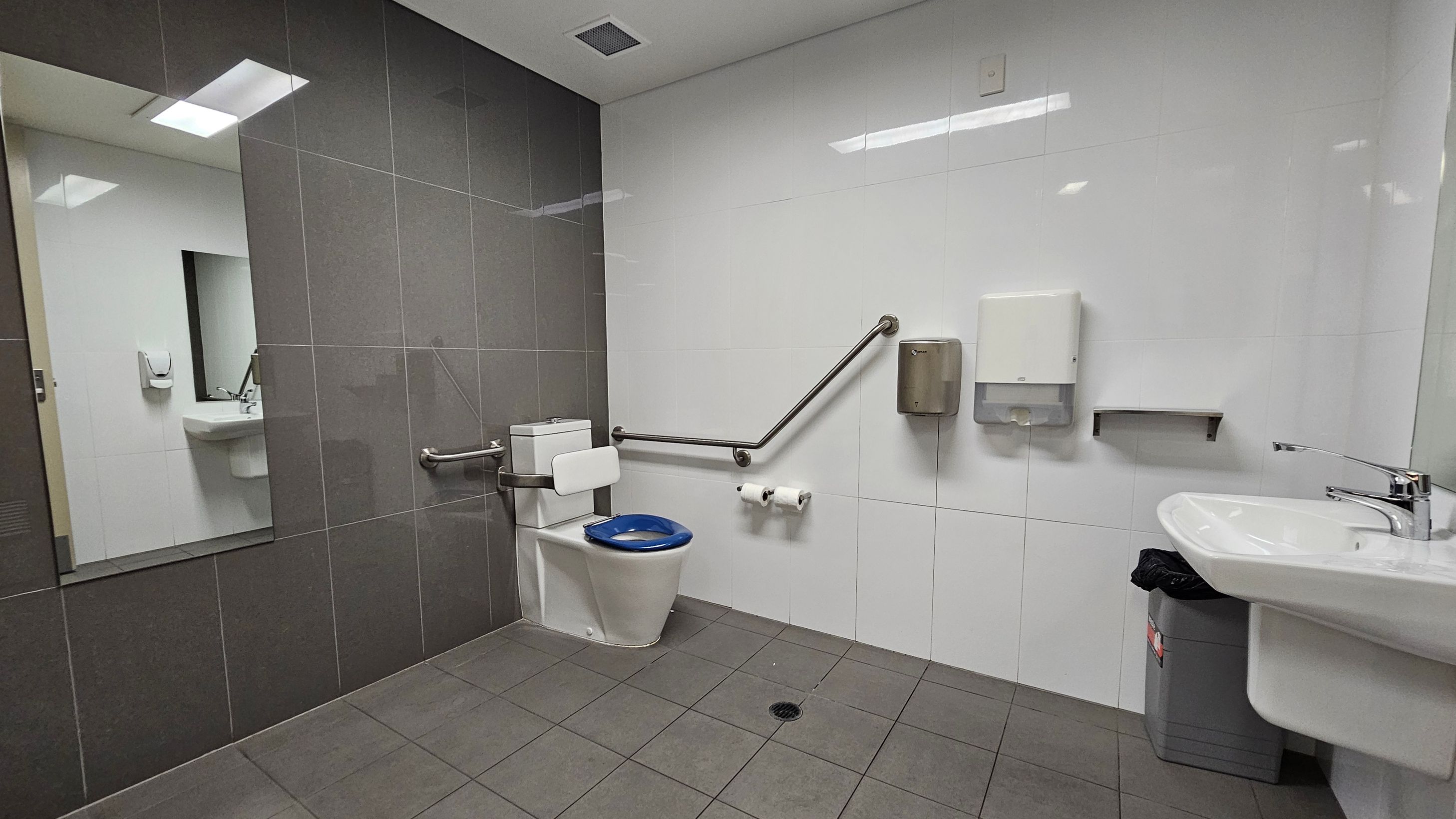 A public restroom with grey floor tiles and a combination of grey and white wall tiles. On the left, a large mirror reflects a sink and soap dispenser. In the centre, a toilet with a blue seat is equipped with support rails. Above the toilet, there are two toilet paper rolls, a hand dryer, and a paper towel dispenser. On the right, a white sink with a chrome tap is mounted on the wall, with a grey bin underneath. The ceiling is white with recessed lighting.