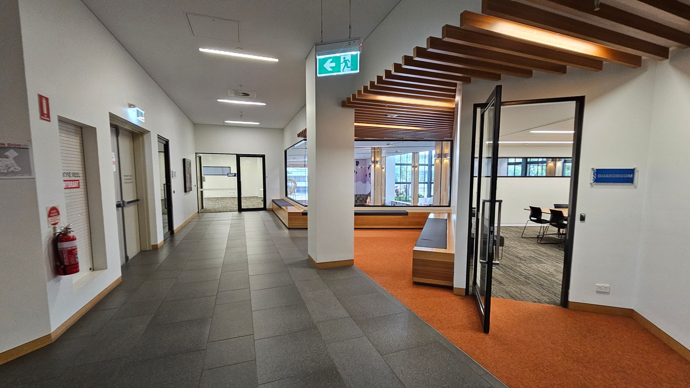 A hallway with grey tiled flooring and a white ceiling. On the left, there are doors and a fire extinguisher mounted on the wall. In the centre, a glass door leads to another area with a visible seating area and large windows. On the right, an open door with a "Boardroom" sign reveals a room with chairs and a carpeted floor. The ceiling features wooden slats above the boardroom entrance, and there is an emergency exit sign hanging from the ceiling.