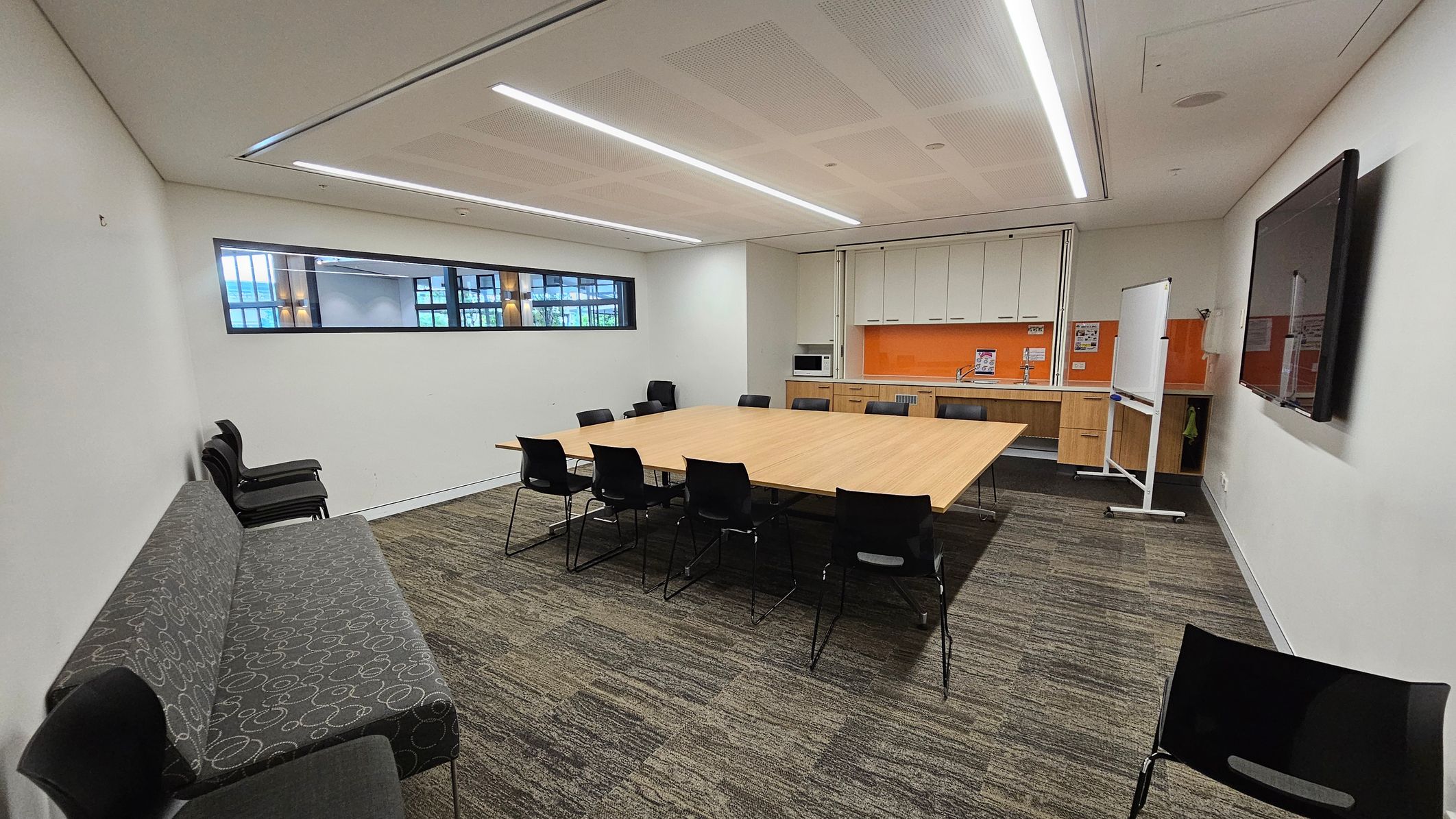 A conference room with a large rectangular wooden table in the centre, surrounded by black chairs. On the left, a grey patterned sofa and stacked chairs are against the wall. The right side features a wall-mounted TV and a whiteboard on wheels. The back wall has a kitchenette with orange backsplash, cabinets, and a microwave. A narrow window runs along the upper left wall. The floor is carpeted in a green and brown pattern, and the ceiling has recessed lighting.
