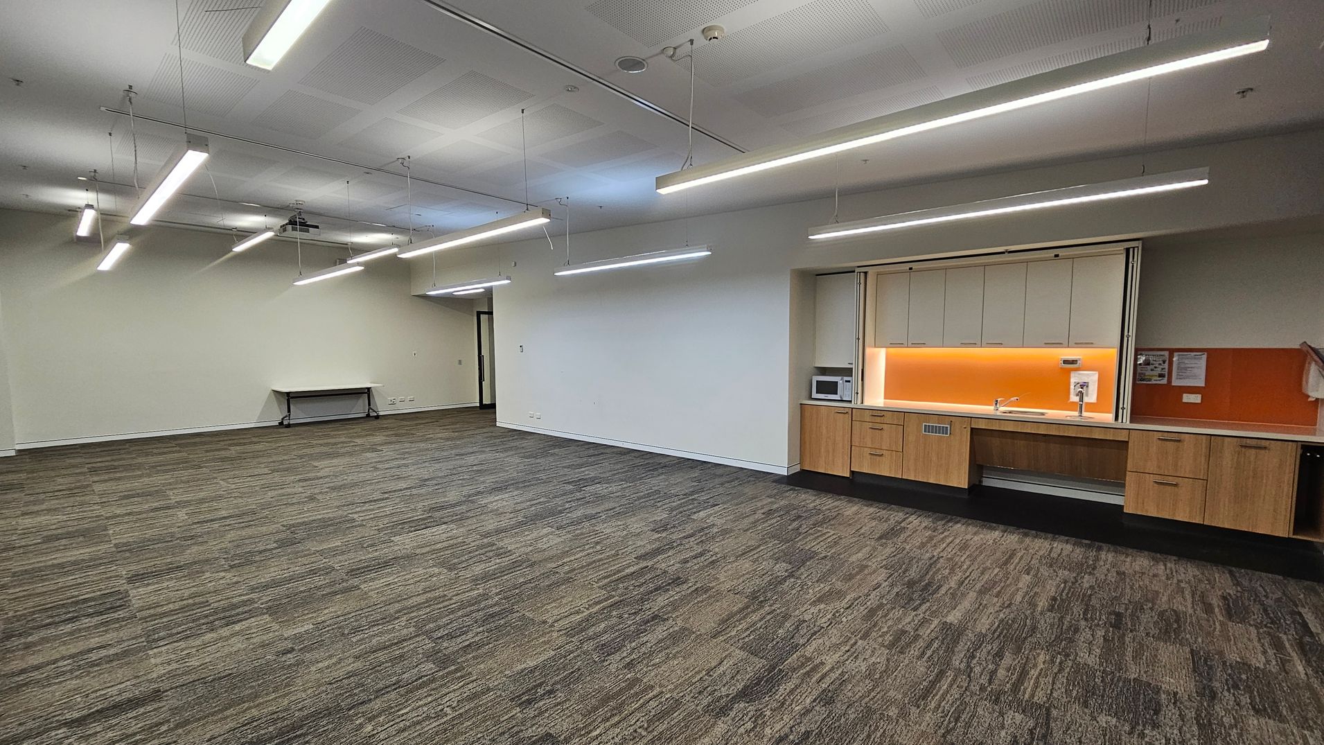 A spacious room with a patterned carpet in shades of green and brown. On the right, there is a kitchenette with wooden cabinets, a microwave, and a sink, set against an orange backsplash. The ceiling features several long, suspended fluorescent lights. On the left, a small table is positioned against a pale green wall. The room has a modern, minimalist design with a focus on functionality.