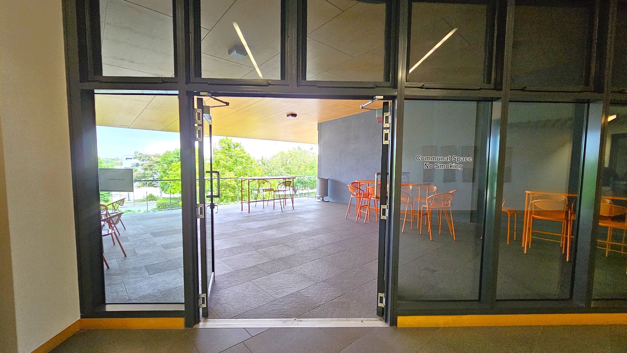 Glass doors open to a communal space with a "No Smoking" sign. Inside, there are orange chairs and tables on a tiled floor. The ceiling is light-coloured with linear lights. To the left, there's a view of greenery through the open area. The right side shows more seating arrangements. The space appears to be a balcony or terrace with a railing visible in the background.