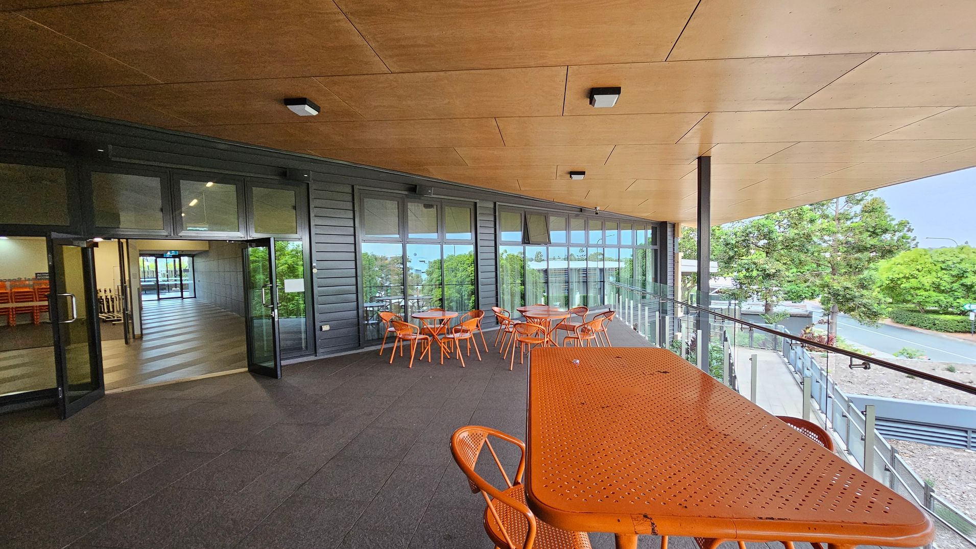 A covered outdoor area with a wooden ceiling and dark flooring. On the left, there are glass doors leading to an indoor space with stacked chairs. In the middle, several orange tables and chairs are arranged near the glass wall. On the right, a long orange table with chairs is positioned near a glass railing overlooking trees and a road.
