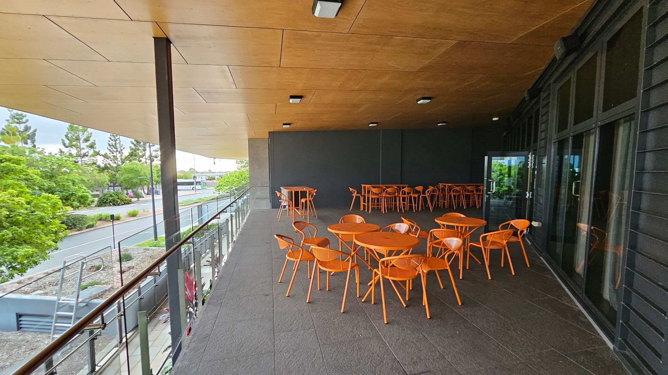 A covered outdoor seating area with orange tables and chairs on a grey tiled floor. On the right, there are large glass doors. The ceiling is wooden with recessed lighting. On the left, a glass railing overlooks a street lined with trees and greenery.
