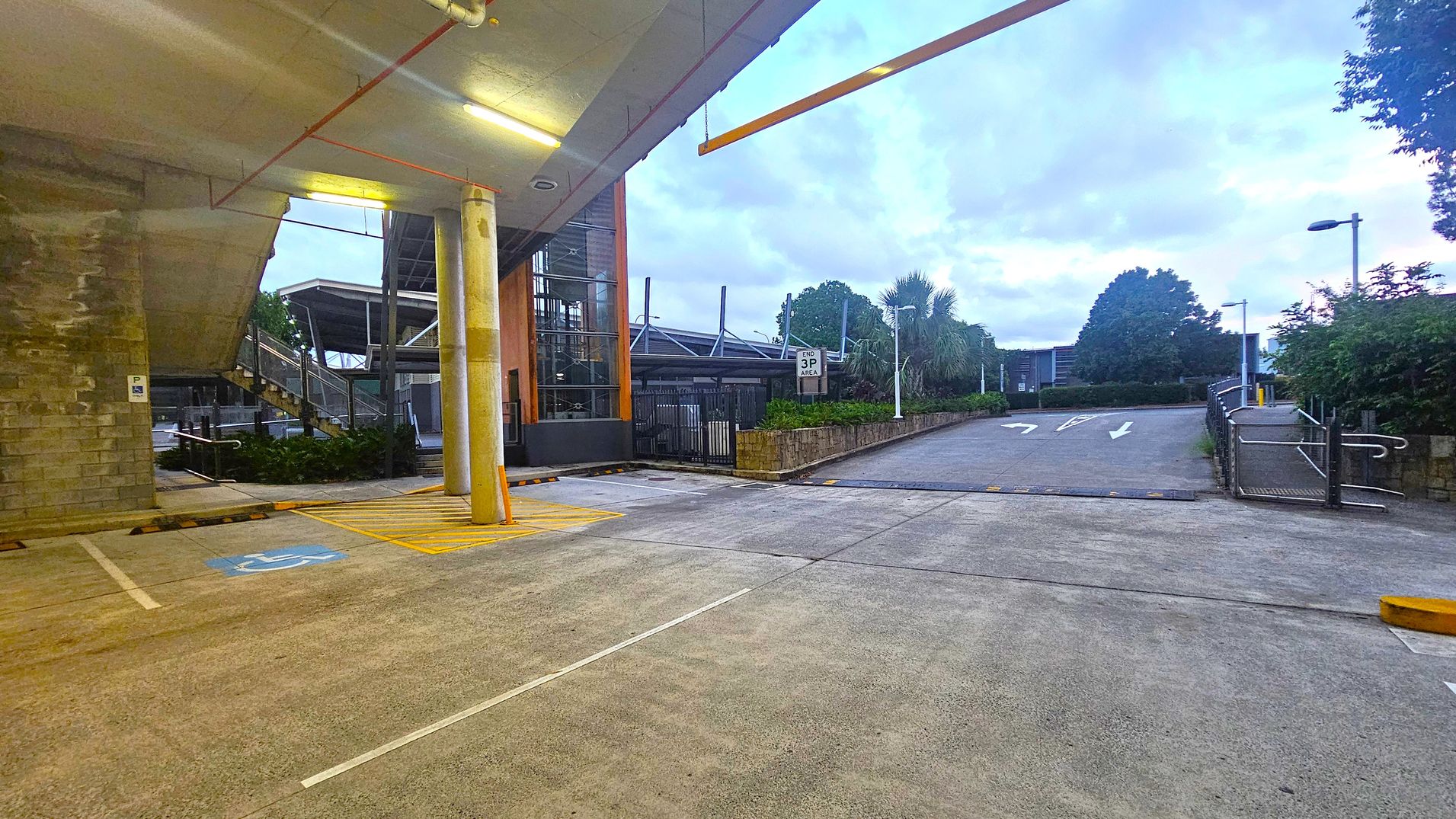 A parking area with a concrete floor and a ceiling with fluorescent lights. On the left, there's a brick wall and a staircase leading up. A yellow-striped area and a disabled parking spot are visible. In the centre, a tall pillar supports the structure. To the right, a driveway with white arrows leads upwards, bordered by greenery and a metal railing. A sign reads "END 3P AREA." Trees and shrubs are in the background.