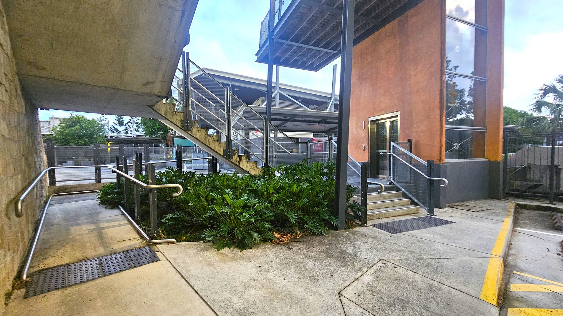 A concrete pathway with metal railings leads to a building entrance on the right, featuring a glass and orange facade. A staircase with metal railings ascends beside the building. Dense green foliage is situated between the path and the stairs. The background includes a fence and trees, with a partially visible structure. The ground is concrete with yellow markings, and the ceiling is not visible.
