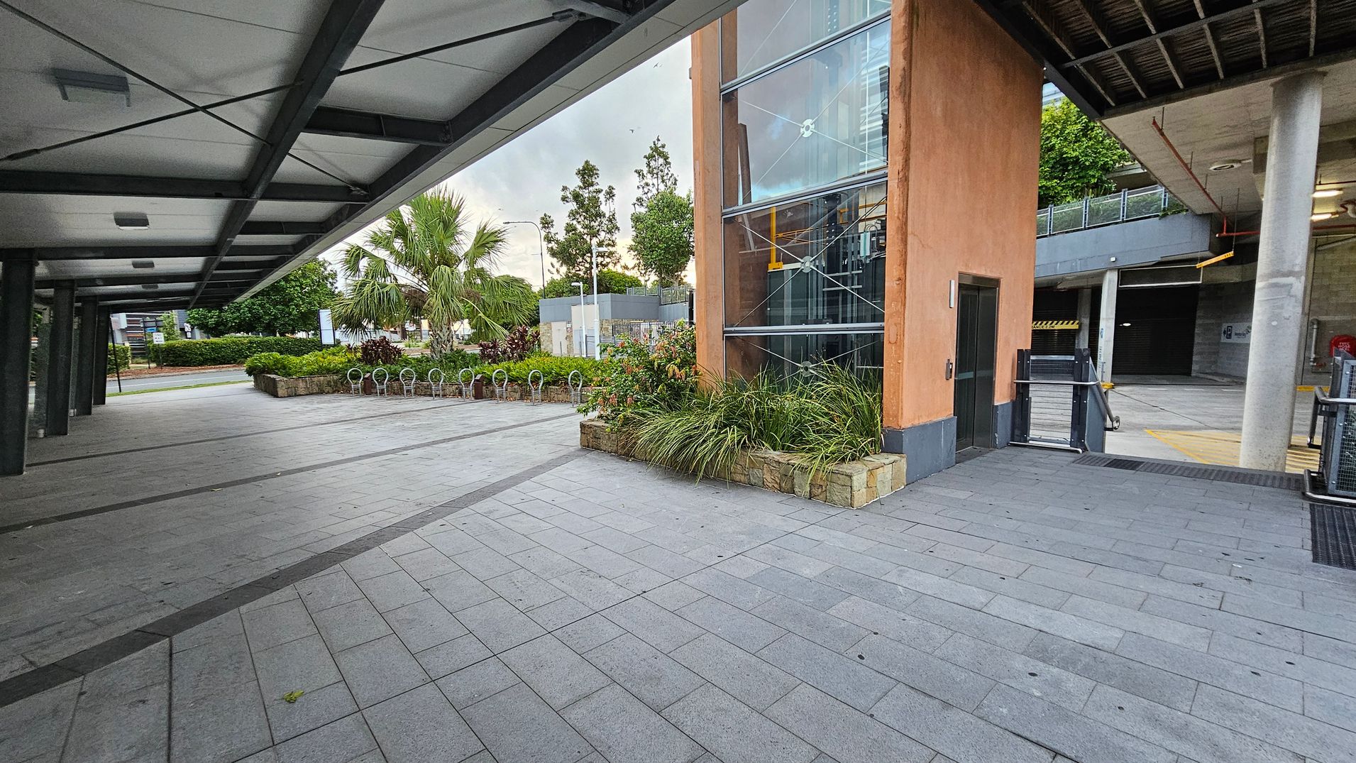 A paved walkway with a covered ceiling on the left, featuring metal beams. On the right, there's a building with a glass facade and an orange wall, surrounded by planters with green plants. In the background, there are trees and a bicycle rack. The area appears open and spacious, with a mix of natural and urban elements.