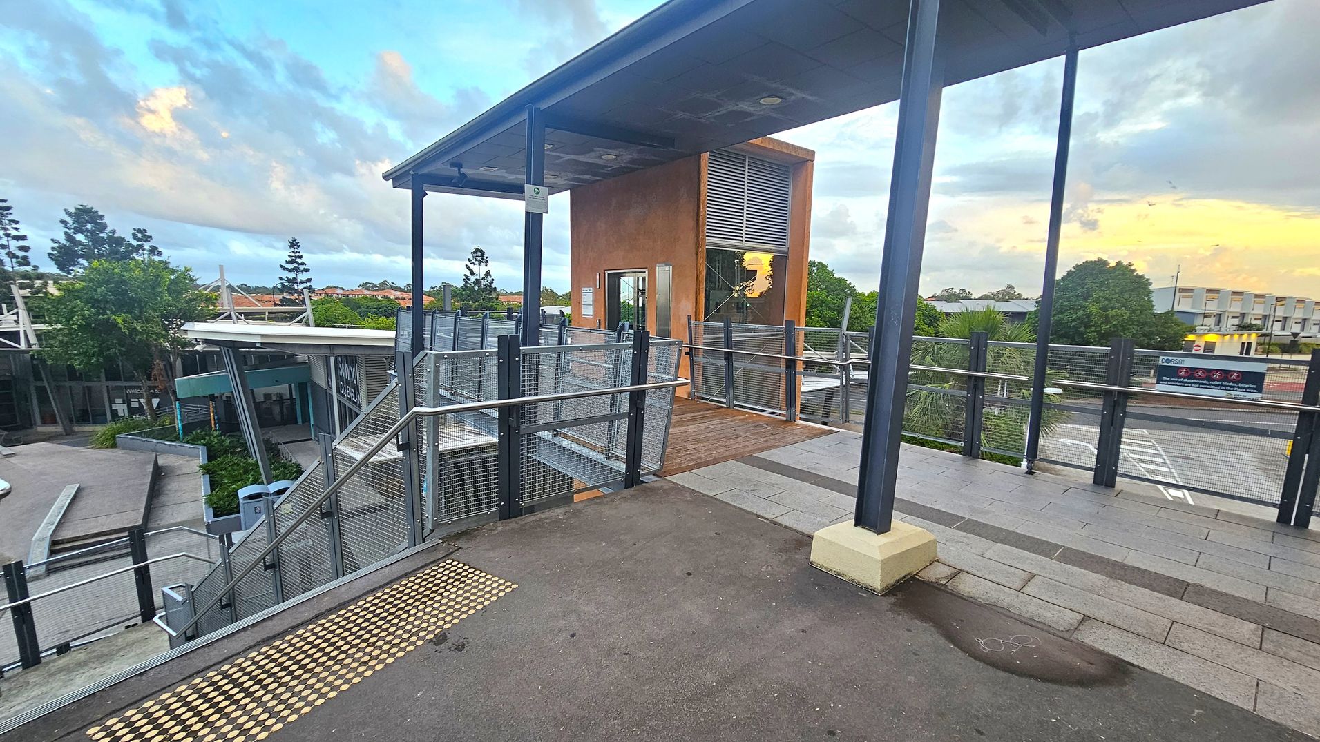 A modern outdoor walkway with a metal railing and a ramp leading to a small building with a glass door and a brown exterior. The floor is a mix of concrete and tiles, with tactile paving for accessibility. On the left, there are trees and a lower level with more structures. On the right, a road and more greenery are visible. The ceiling is a simple metal structure supported by dark columns.