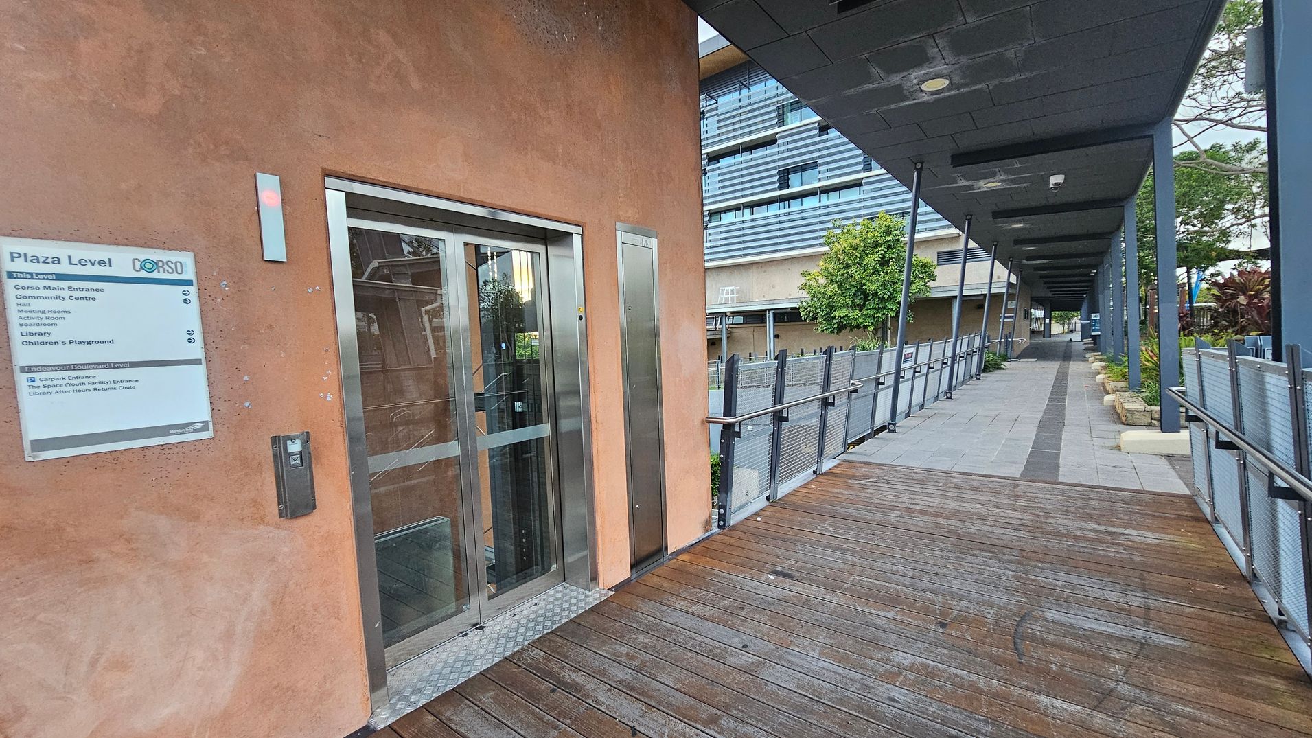 A wooden walkway leads to a glass lift on the left, set in a peach-coloured wall. A sign next to the lift lists facilities like a community centre and library. On the right, a long covered pathway with a metal railing and potted plants runs parallel to the building. The ceiling is dark with recessed lighting, and a modern building with horizontal lines is visible in the background.