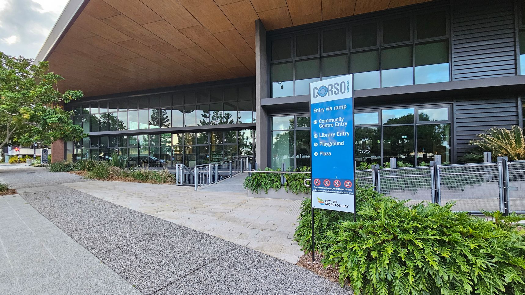 A modern building with large glass windows and a wooden overhang. On the right, a blue sign lists entries for a community centre, library, playground, and plaza. The foreground features a paved walkway and lush green plants. On the left, a tree provides shade. The building's facade includes dark panels and a metal railing along a ramp leading to the entrance.