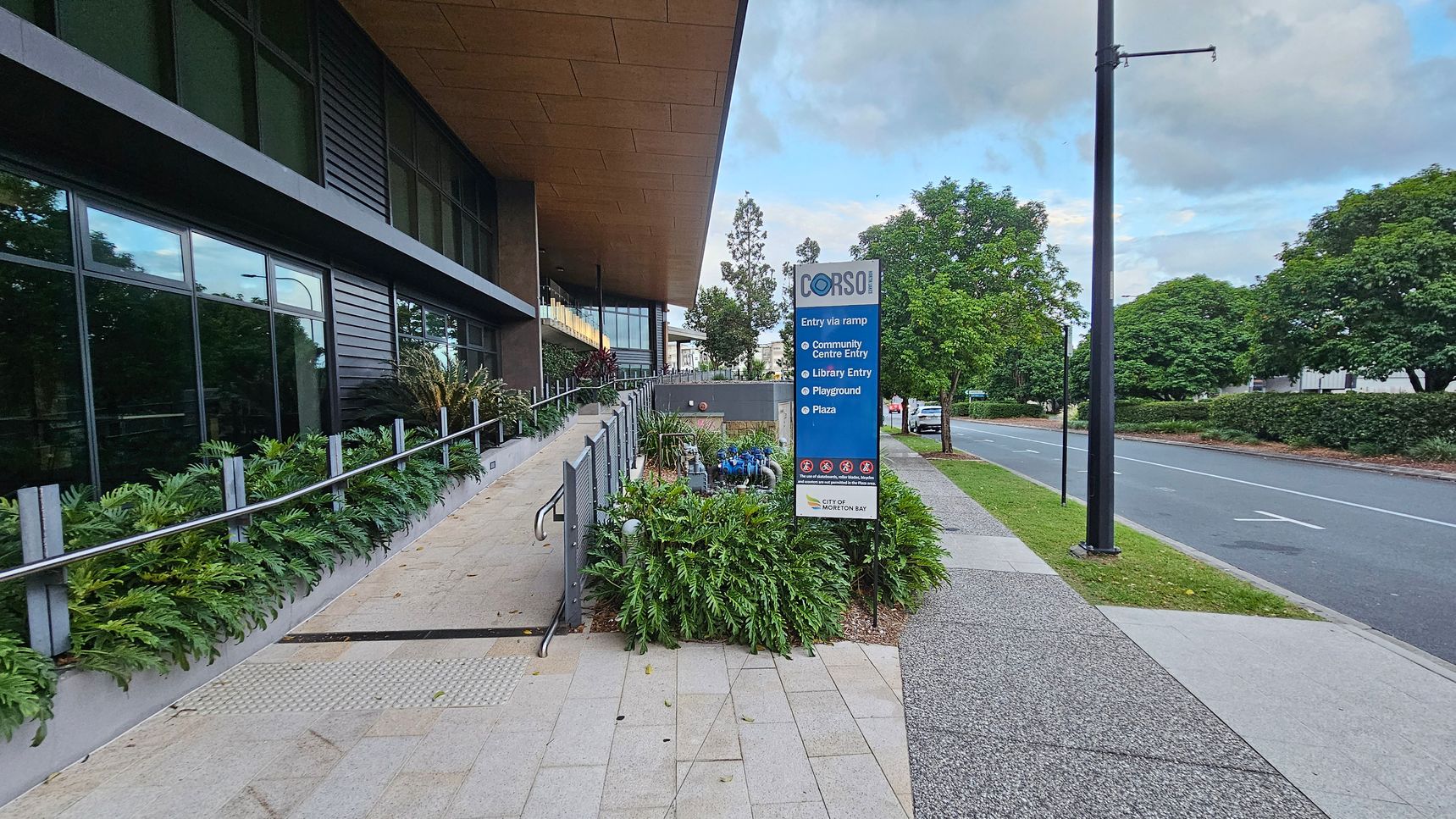 On the left, a modern building with large glass windows and a sloped roof. A ramp with metal railings is lined with lush green plants. In the centre, a blue sign lists various entries and facilities, including a community centre and library. On the right, a road bordered by grass and trees, with a streetlamp and a parked car visible. The pavement is light-coloured and textured.