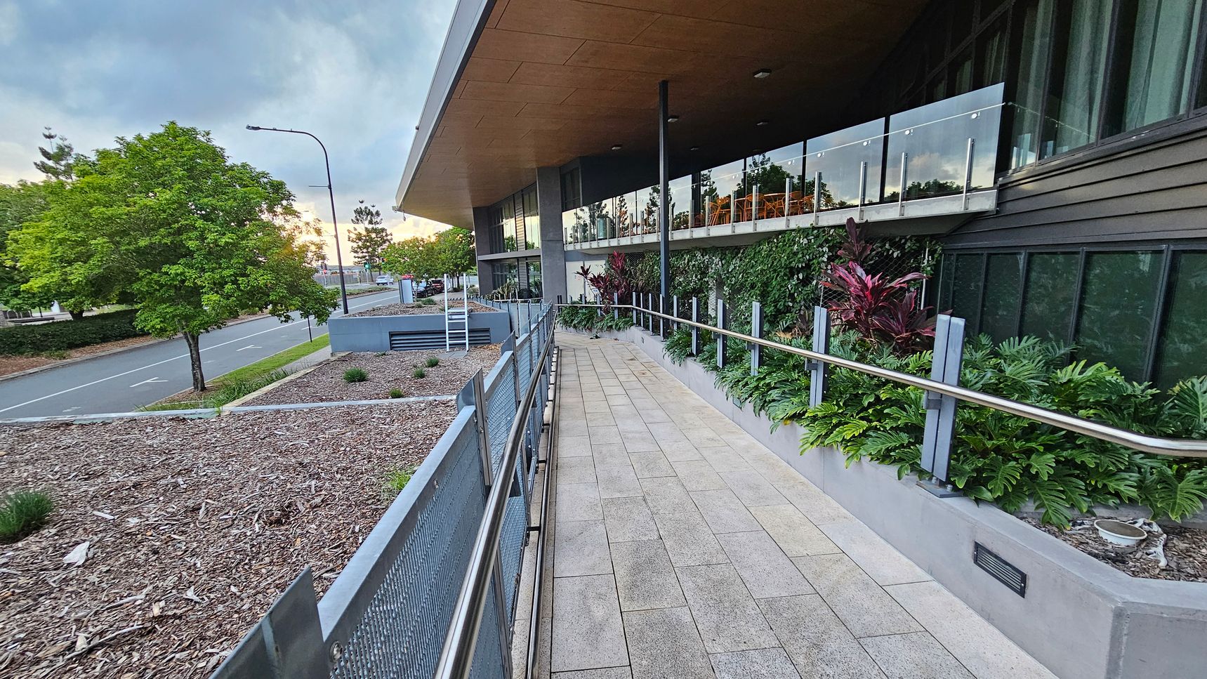A paved walkway with metal railings runs alongside a modern building on the right, featuring large glass windows and a wooden overhang. Lush green plants line the path. On the left, a road with a few trees and a streetlamp is visible. The ground on the left is covered with mulch and small plants. The ceiling of the overhang is wooden, and the floor is tiled.