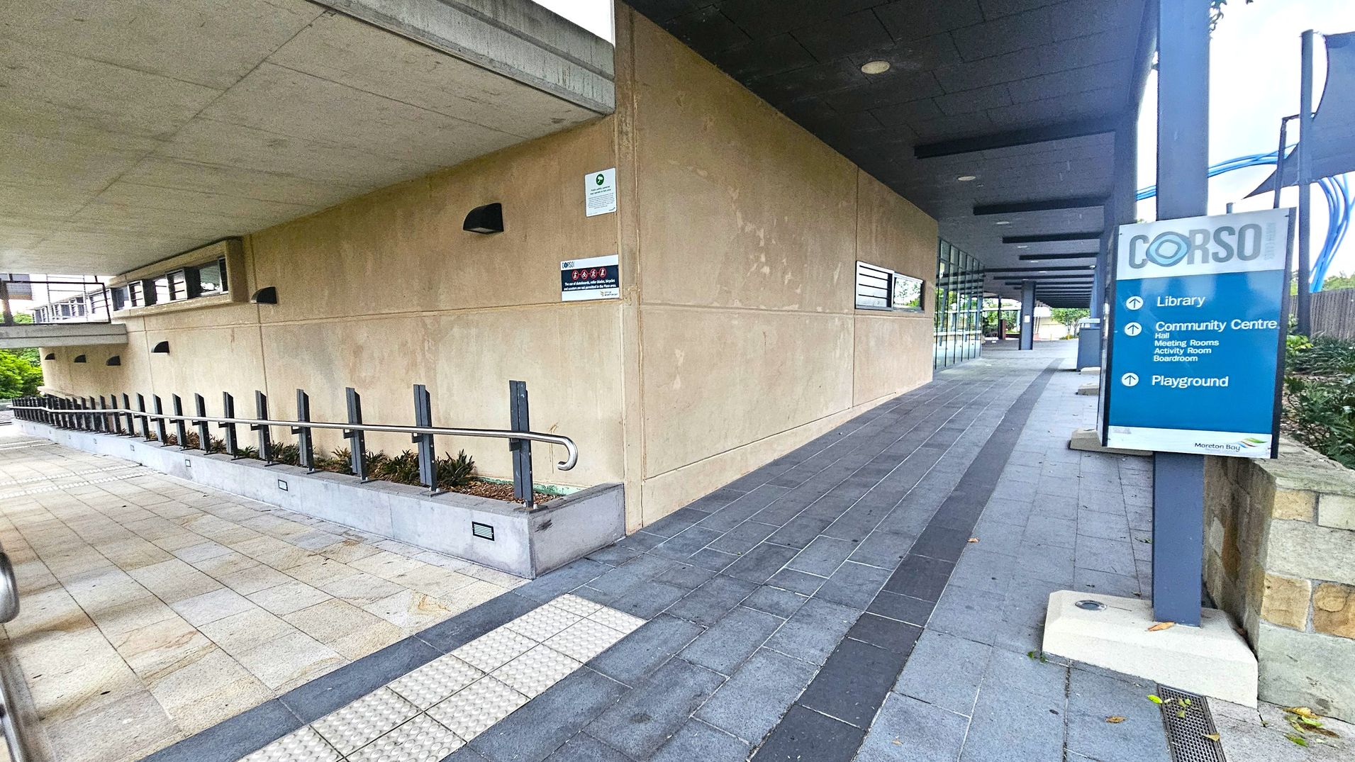 A walkway with a beige wall on the left featuring small windows and black wall lights. A metal railing runs alongside a row of small plants. On the right, a blue sign lists directions to a library, community centre, and playground. The path is paved with grey tiles, and the ceiling is dark with recessed lighting. In the background, there are more plants and a continuation of the building structure.