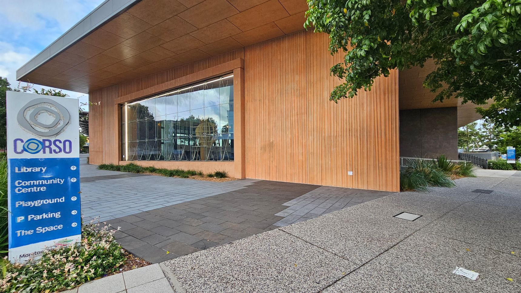 A modern building with a wooden exterior and large glass windows. On the left, a sign reads "Corso" with directions to a library, community centre, playground, parking, and "The Space." The ground is paved with grey and black tiles, and there are small plants near the sign. A tree with green leaves is on the right, partially shading the area. The ceiling is wooden, matching the building's exterior.