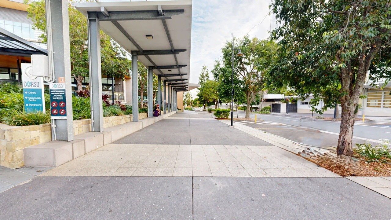 A paved walkway with a covered area on the left, supported by metal columns. A sign with text and symbols is attached to one of the columns. To the right, there are trees and a street with a few parked cars. The background features more trees and buildings. The ground is a mix of grey and beige paving, and the ceiling of the covered area is flat with recessed lighting.