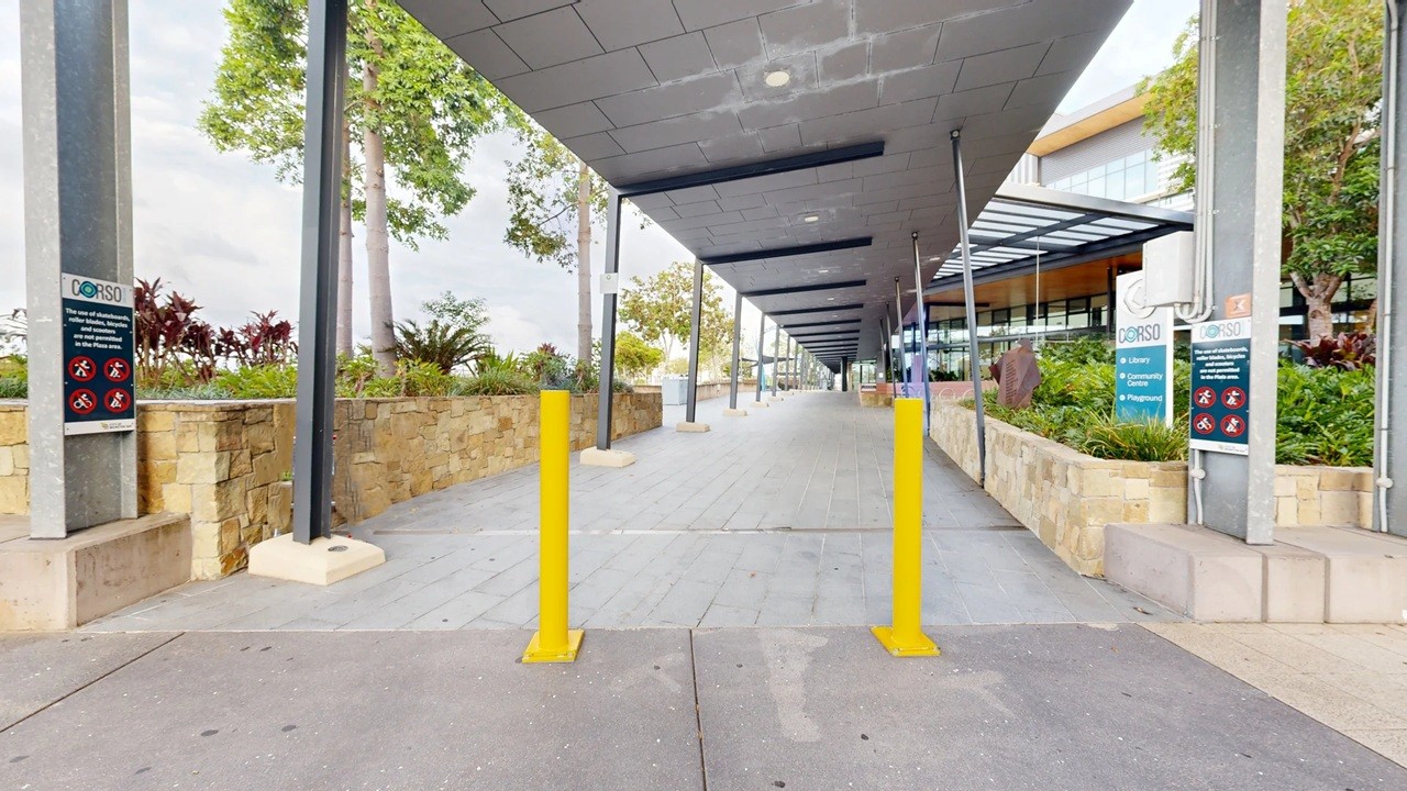 A covered walkway with a grey ceiling and concrete floor. Two yellow bollards are positioned in the foreground. On the left, a stone wall with plants and a sign attached to a metal post. On the right, a similar stone wall with greenery and a sign. The background features a building with glass windows and a metal roof. Trees and more plants are visible along the walkway.