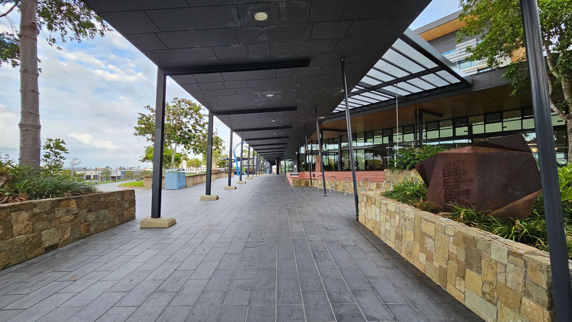 A covered walkway with a dark tiled ceiling supported by black poles runs through the centre. The floor is paved with grey tiles. On the right, a modern building with large glass windows and a metal awning is visible. A stone wall with plants and a geometric sculpture is also on the right. On the left, there are trees and a stone wall with greenery. In the distance, there are more trees and structures.