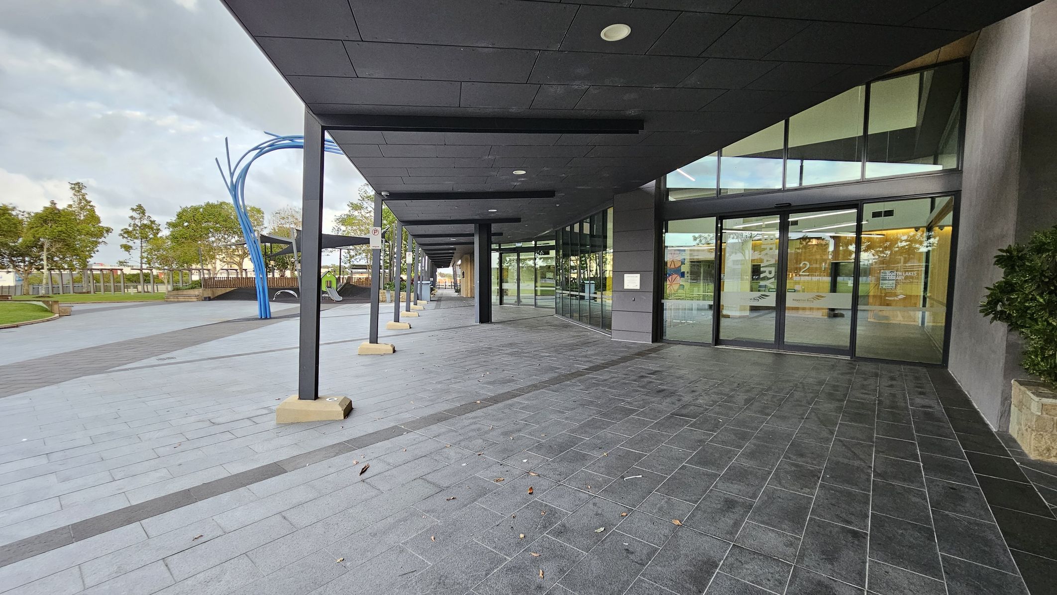 A modern building entrance with large glass doors on the right, reflecting the surroundings. The floor is paved with dark grey tiles. On the left, there is an open area with a few trees and a blue sculptural structure. The ceiling is dark with recessed lighting. The area is spacious and clean, with a mix of natural and architectural elements.