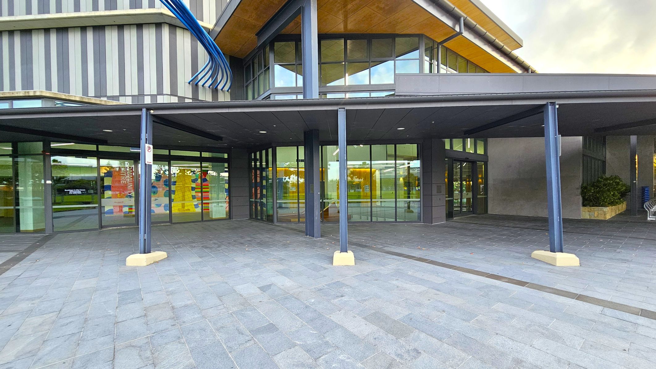 The image shows the entrance of a modern building with large glass windows and a canopy supported by blue pillars. The facade features vertical stripes in various shades of grey. On the left, colourful patterns are visible through the windows. The floor is paved with grey tiles, and there is a small plant in a stone planter on the right. The ceiling of the canopy is smooth and dark, contrasting with the wooden underside of the building's roof.