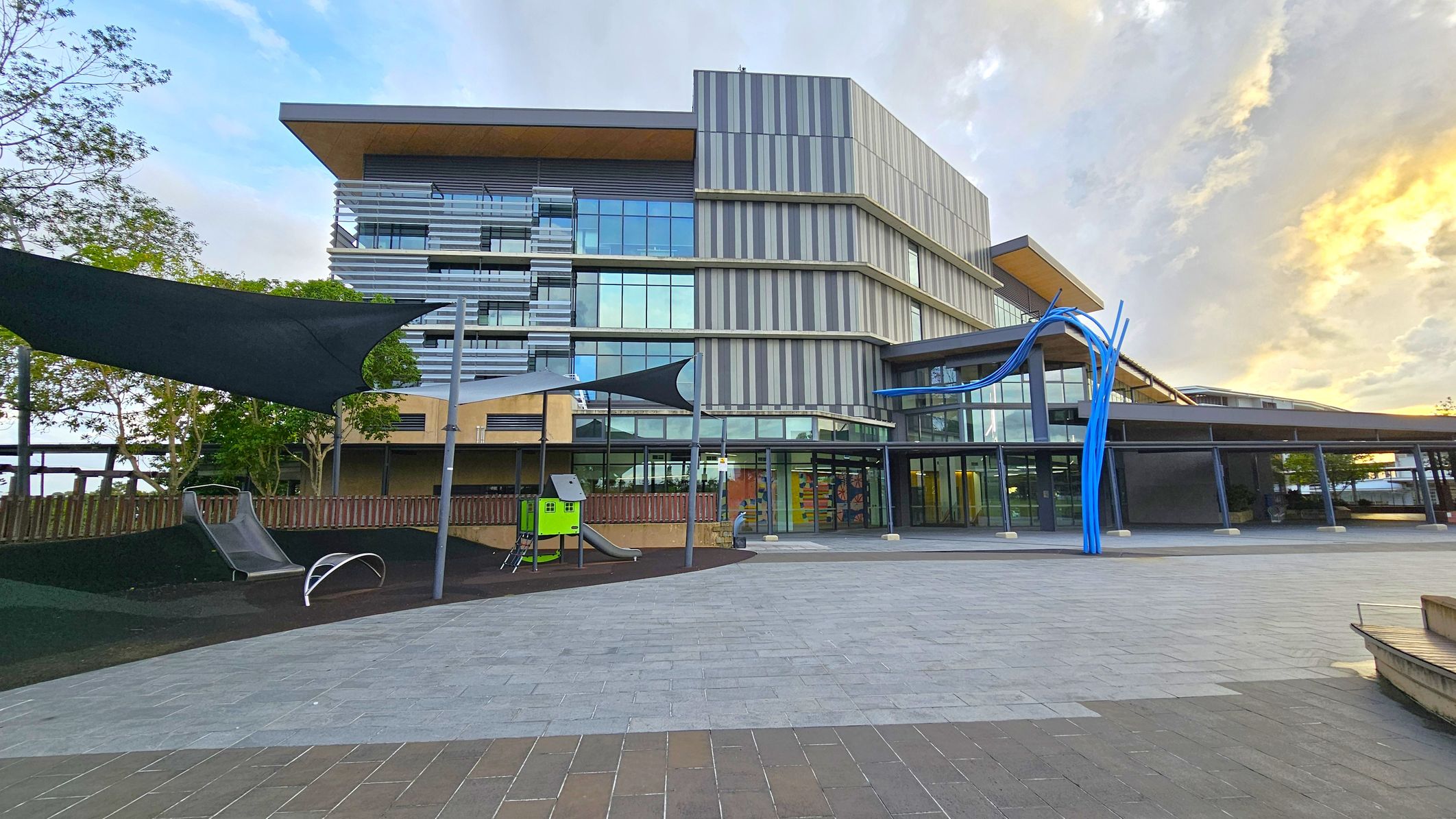 A modern building with a multi-storey design featuring glass and striped panels. On the left, there's a playground area with a slide and a green play structure, shaded by dark triangular canopies. The foreground has a paved area with grey and brown tiles. On the right, a blue abstract sculpture stands near the building's entrance, which has large glass doors. Trees are visible in the background, adding greenery to the scene.