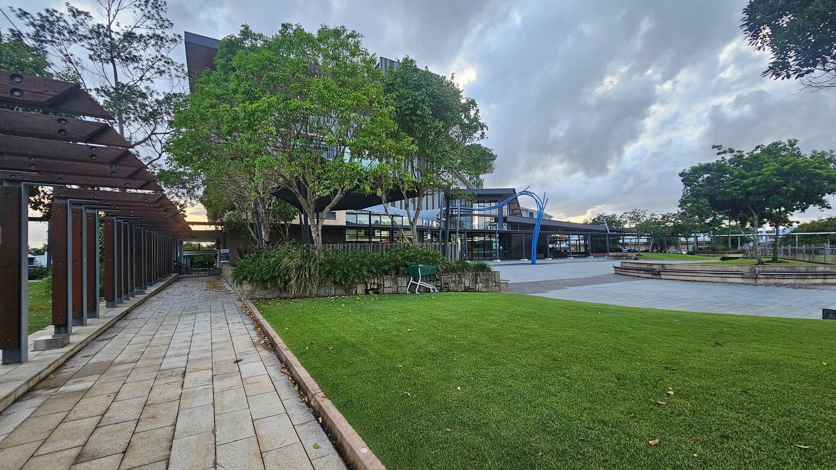 A paved walkway on the left is lined with a wooden pergola and leads towards a building partially obscured by trees. In the centre, a grassy area with a shopping trolley is bordered by a low stone wall. The building features large glass windows and a modern design. On the right, a plaza with grey tiles and a few trees is visible, along with a blue sculptural element. The sky is overcast with grey clouds.