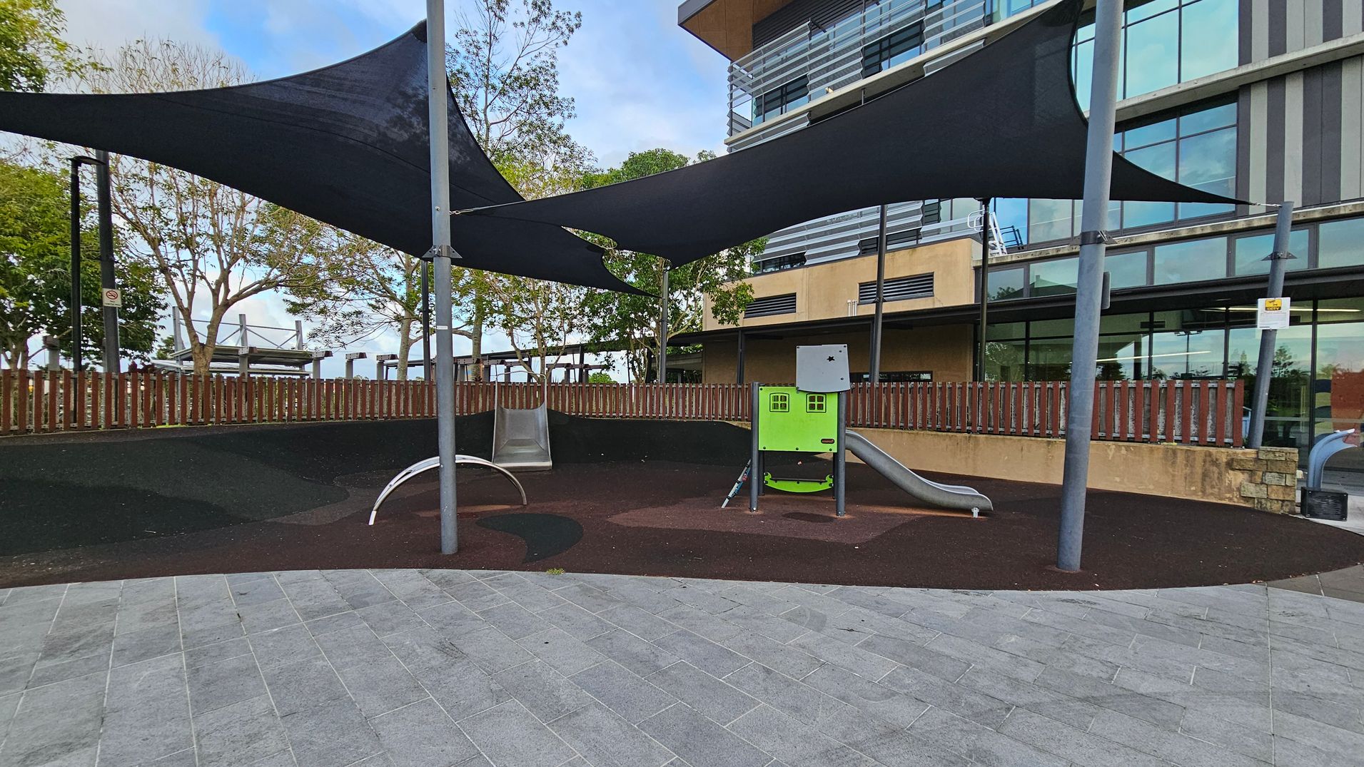 A playground area with a grey and purple rubber surface. On the left, there's a metal slide and a curved climbing structure. In the centre, a small green playhouse with a slide is visible. Overhead, black shade sails are supported by metal poles. In the background, a modern building with large glass windows and a wooden fence is visible. Trees are scattered around the area. The foreground features a grey tiled floor.