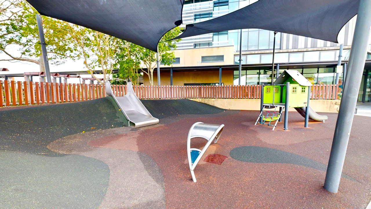 A playground with a rubberised surface. On the left, there's a metal slide. In the centre, a curved climbing structure is visible. On the right, a small playhouse with a slide and climbing net. The background features a wooden fence and a modern building with large windows. Overhead, there are dark shade sails providing cover. Trees are visible behind the fence.
